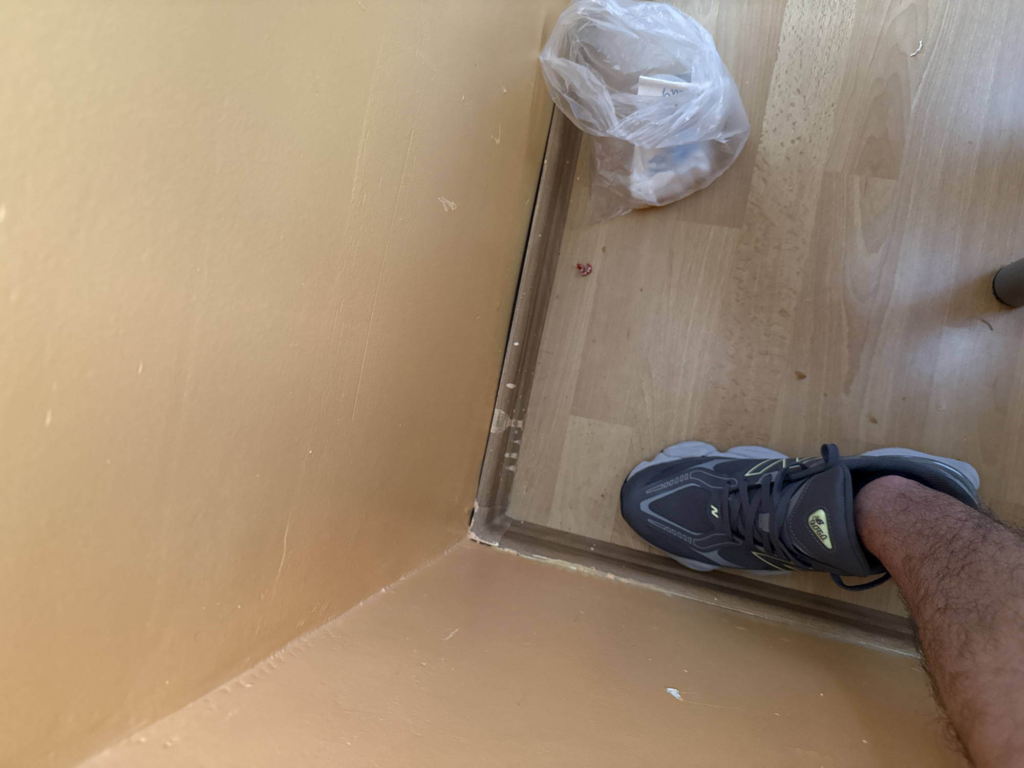 The image displays an indoor corner, looking down at a light tan-colored wall and a light wooden floor. A dark brown transition strip runs along the base of the wall where it meets the floor, with some light-colored residue, possibly caulk, visible.
In the bottom right, a person's lower leg, covered in hair, is visible, wearing a dark grey New Balance 9060 sneaker with yellow accents. The New Balance brand name and "9060" model number are clearly visible on the shoe's tongue.
On the wooden floor, near the wall in the upper right, rests a clear plastic bag containing some white or light-colored items, possibly a small cloth or tissue. Faint, handwritten text resembling "6h" is barely discernible on a label inside the bag. To the far right, a dark grey cylindrical object, likely a furniture leg, is partially visible. The wall has some minor scuff marks and textural variations, while the wooden floor shows distinct grain patterns. The scene depicts a casual interior setting.