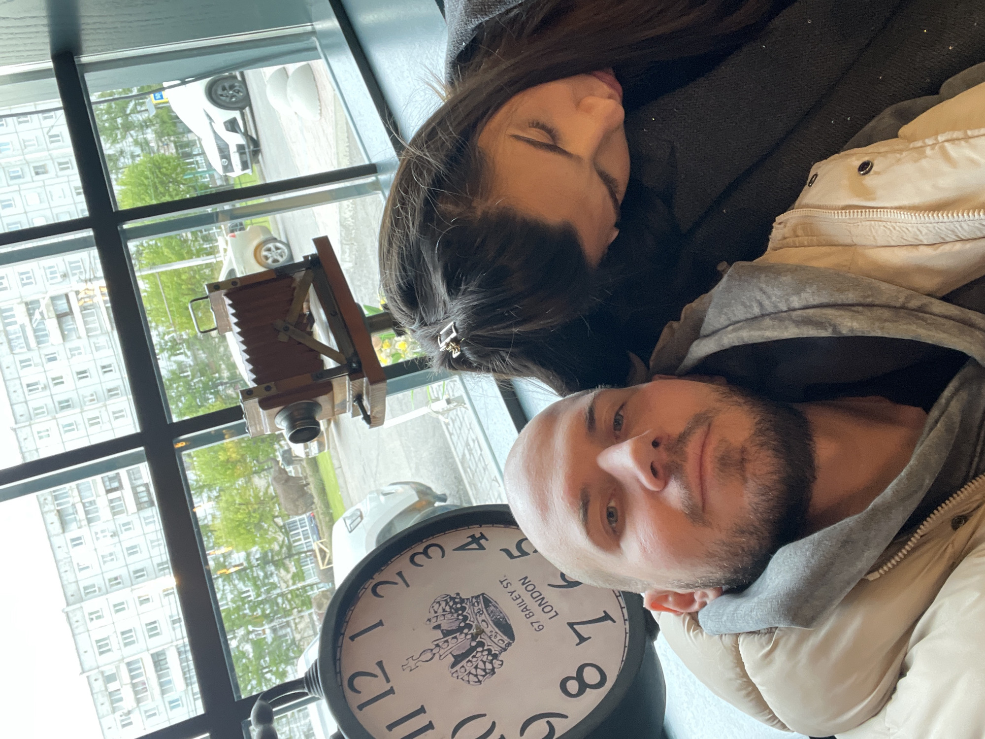Two people are resting, a woman sleeping and a bald man with a beard looking at the camera. Behind them is a window showing a view of apartment buildings and greenery. A vintage camera and a large clock with the inscription "67 Bailey St LONDON" are also visible in the foreground. The location is Krasnoyarsk, Russia.
