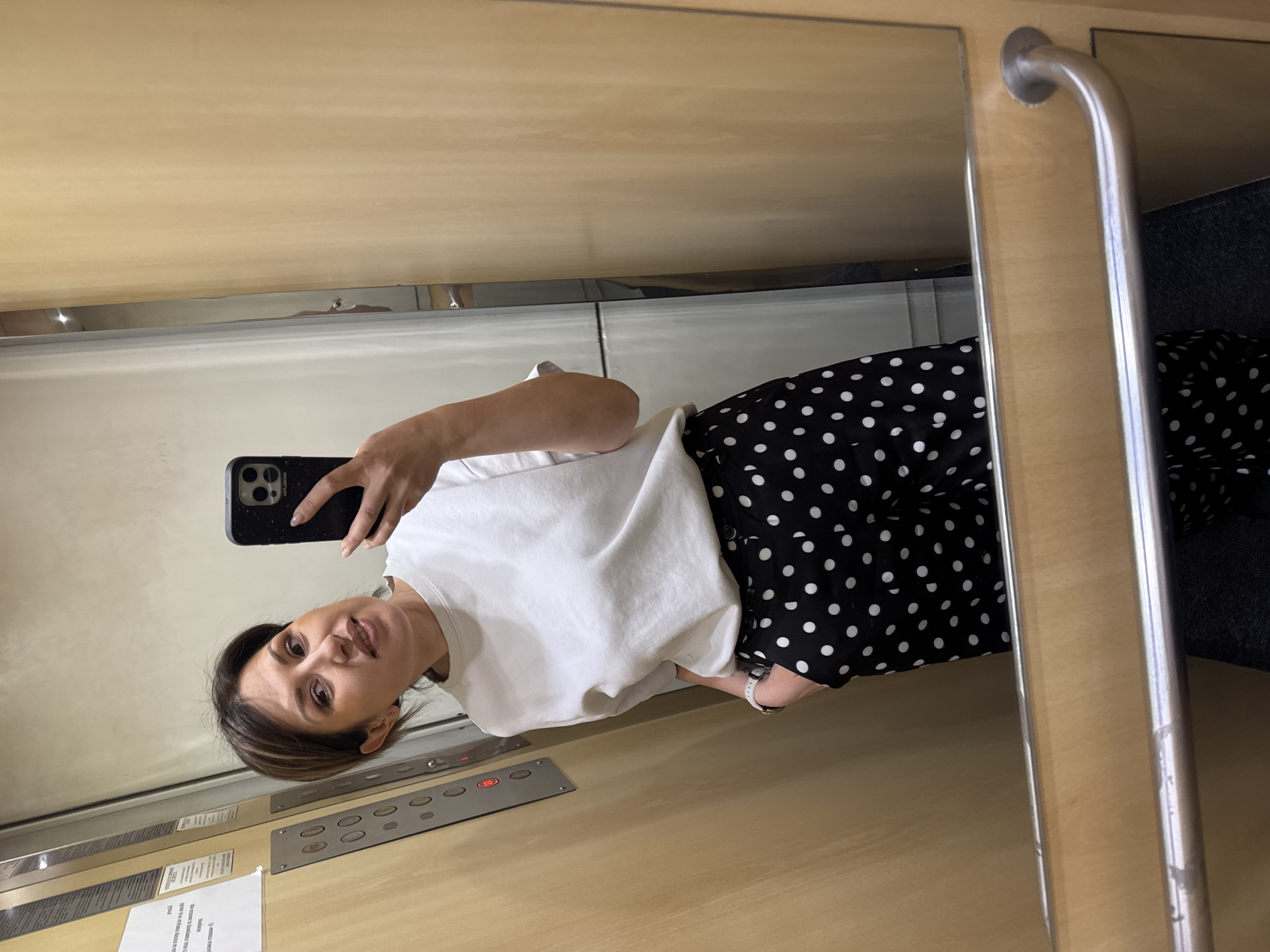 A woman is taking a selfie in a mirror. She is wearing a white t-shirt and a black and white polka dot skirt. She is holding a black smartphone. The background suggests she is in an elevator, possibly in Sofia, Bulgaria. Elevator buttons are visible on the wall, along with a handle. There's a piece of paper with text affixed to the wall.