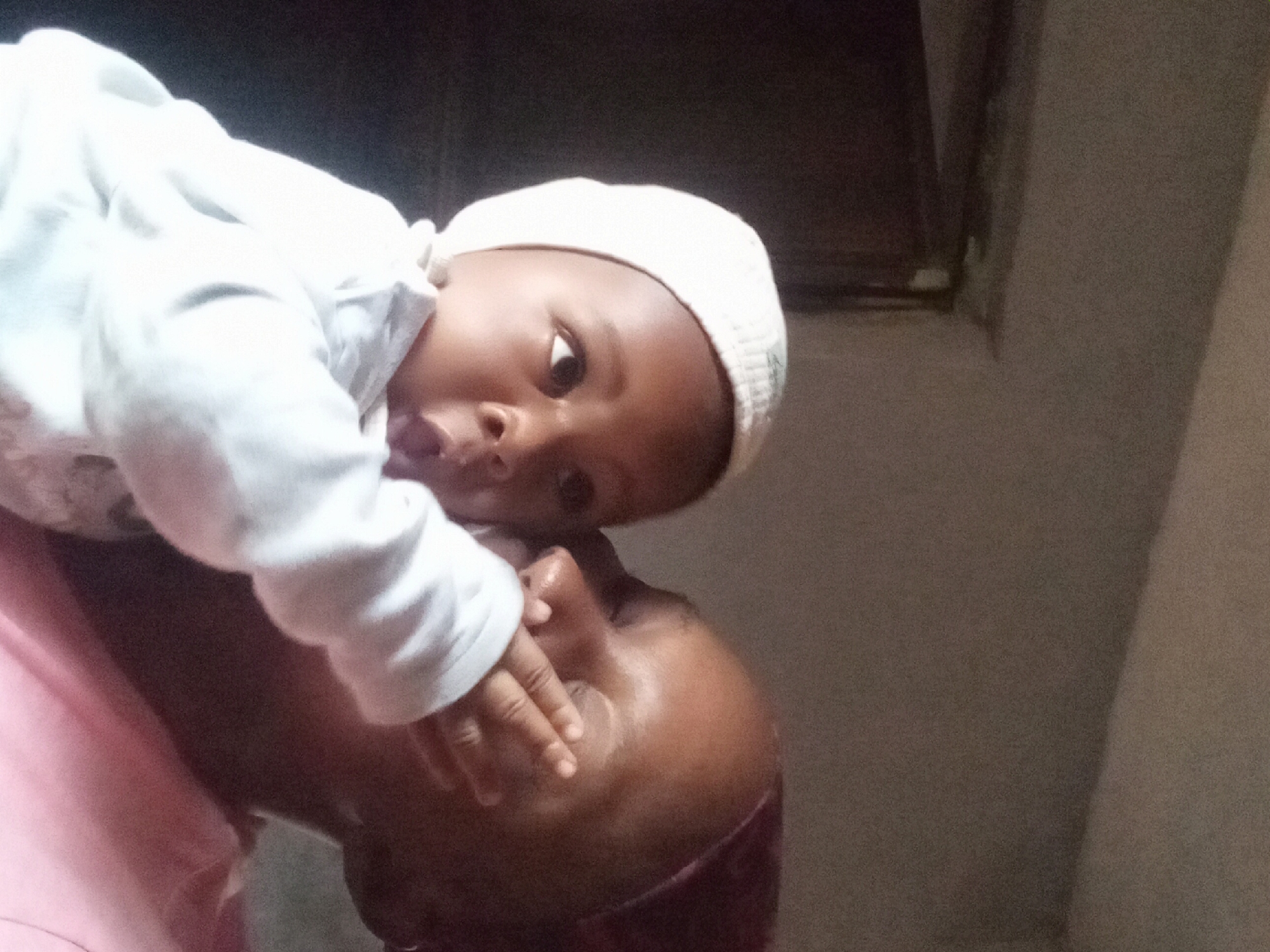 A baby wearing a white cap and a light-colored sweatshirt is held by an adult. The adult is wearing a pink shirt. The interaction between the baby and the adult suggests affection. The background is out of focus, revealing a wall and a dark area. The location is Sabon Tasha, Nigeria.