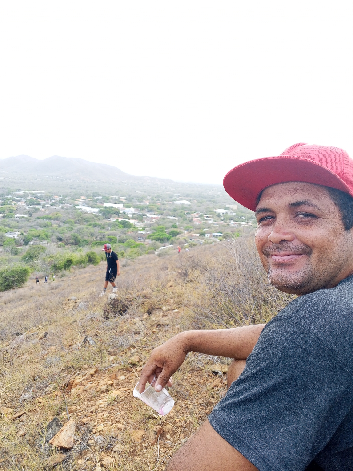 A medium-range outdoor shot depicts a man with a red baseball cap sitting on a hillside, holding a clear plastic cup. The man has dark hair, brown skin, and a slight smile. He is wearing a gray t-shirt. The hillside is dry and covered with sparse vegetation. In the background, there is a valley containing a town with scattered buildings and trees. Beyond the town, a mountain range fades into the hazy sky. A person is visible walking up the hill, wearing a red cap, black shirt, and black shorts. Other distant people are also visible on the hillside. The exact location in the city of Unknown, Unknown is difficult to determine based on the limited information in the image.