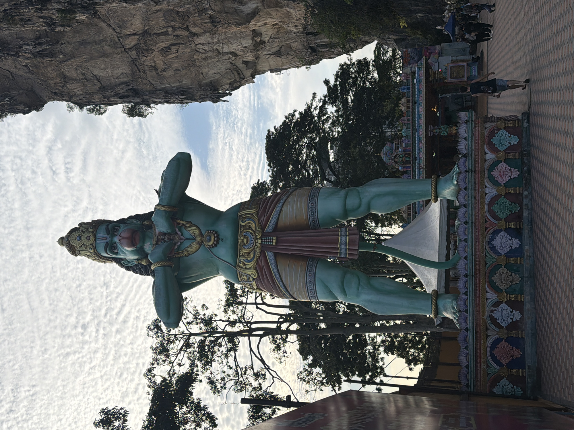 A colossal, teal-skinned statue of a monkey-like deity, adorned with a golden crown, intricate jewelry, and a colorful loincloth, stands prominently in the foreground. The statue's hands are clasped in front of its chest, and its tail curves behind it. It is positioned on a decorative, multi-colored platform. Behind the statue, a large, sheer rock face dominates the right side of the frame. To the left and behind the statue, lush green trees are visible against a cloudy sky. A traditional temple structure with colorful roofs and intricate carvings is situated in the mid-ground, to the right of the statue. Several people are walking on a patterned tiled walkway in front of the temple. The scene appears to be outdoors, likely at a religious site. The location is the Batu Caves, in the district of Gombak, Selangor, within the Selayang Municipal Council area of Malaysia.