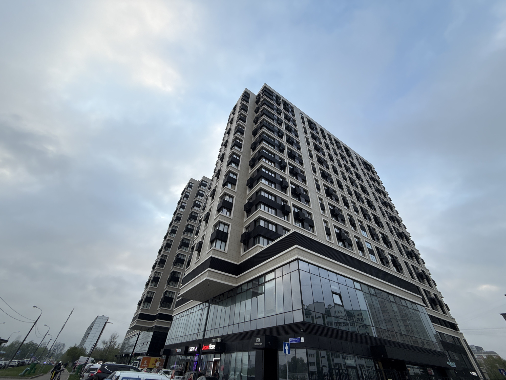 The image shows a modern, multi-story building complex with a facade of light-colored panels and dark-framed windows, set against a cloudy sky. The lower levels of the building feature large glass windows, indicating retail or commercial space. Storefronts such as "OZON" are visible. Cars are parked along the street in front of the building. In the distance, other buildings and streetlights are visible. A street sign that says "Sergeya Eizenshteina" is visible. The location is in Moscow, Russia.