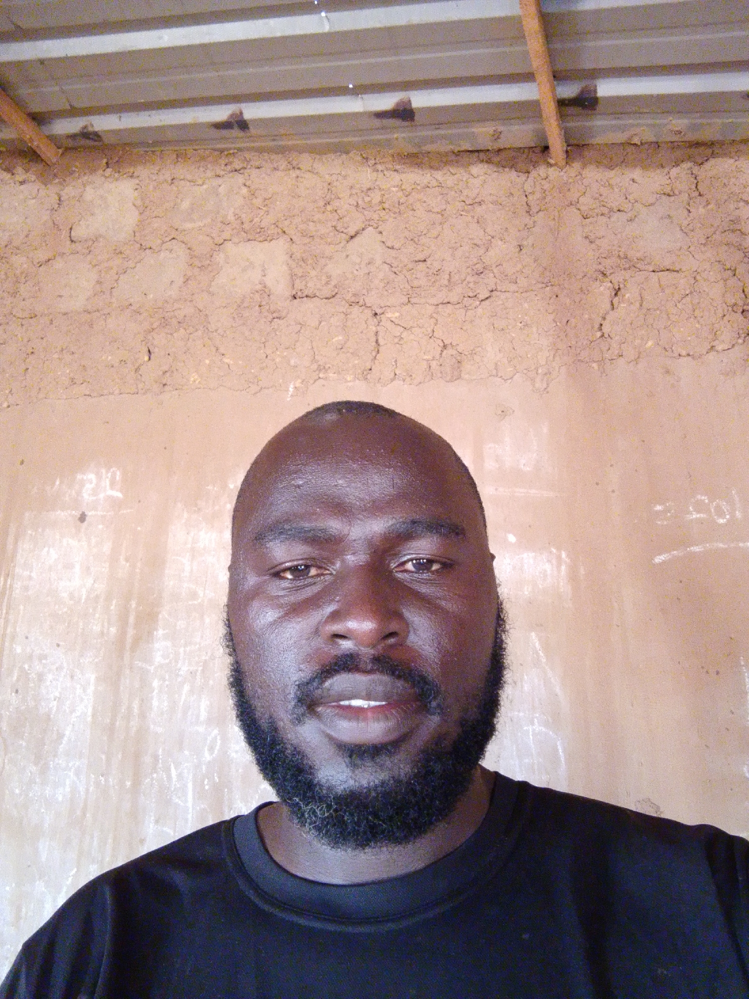 A medium, eye-level shot of a dark-skinned man with a shaved head and a full black beard. He is wearing a black shirt. Behind him is an interior wall constructed of mud brick. Above the mud brick is a corrugated metal roof supported by wooden beams. Writing is visible on the wall. The location is likely Mulke Barikin Sayejo, Nigeria.