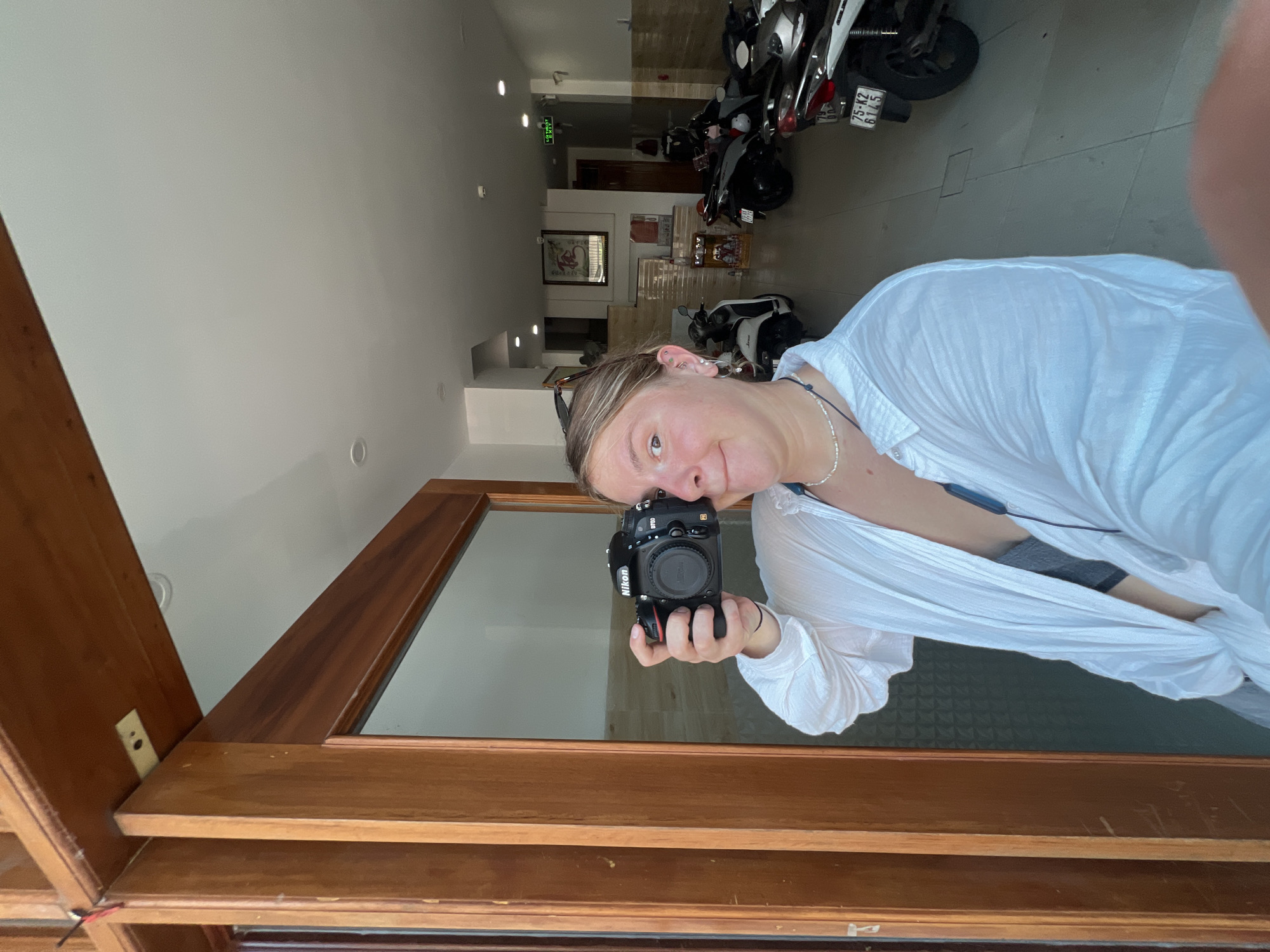 A woman is taking a selfie in a mirror. She is wearing a white shirt and holding a Nikon camera. The reflection shows a hallway in what appears to be a building in Nam Nha Trang Ward, Vietnam. Several scooters are parked in the hallway. There is also artwork on the walls.