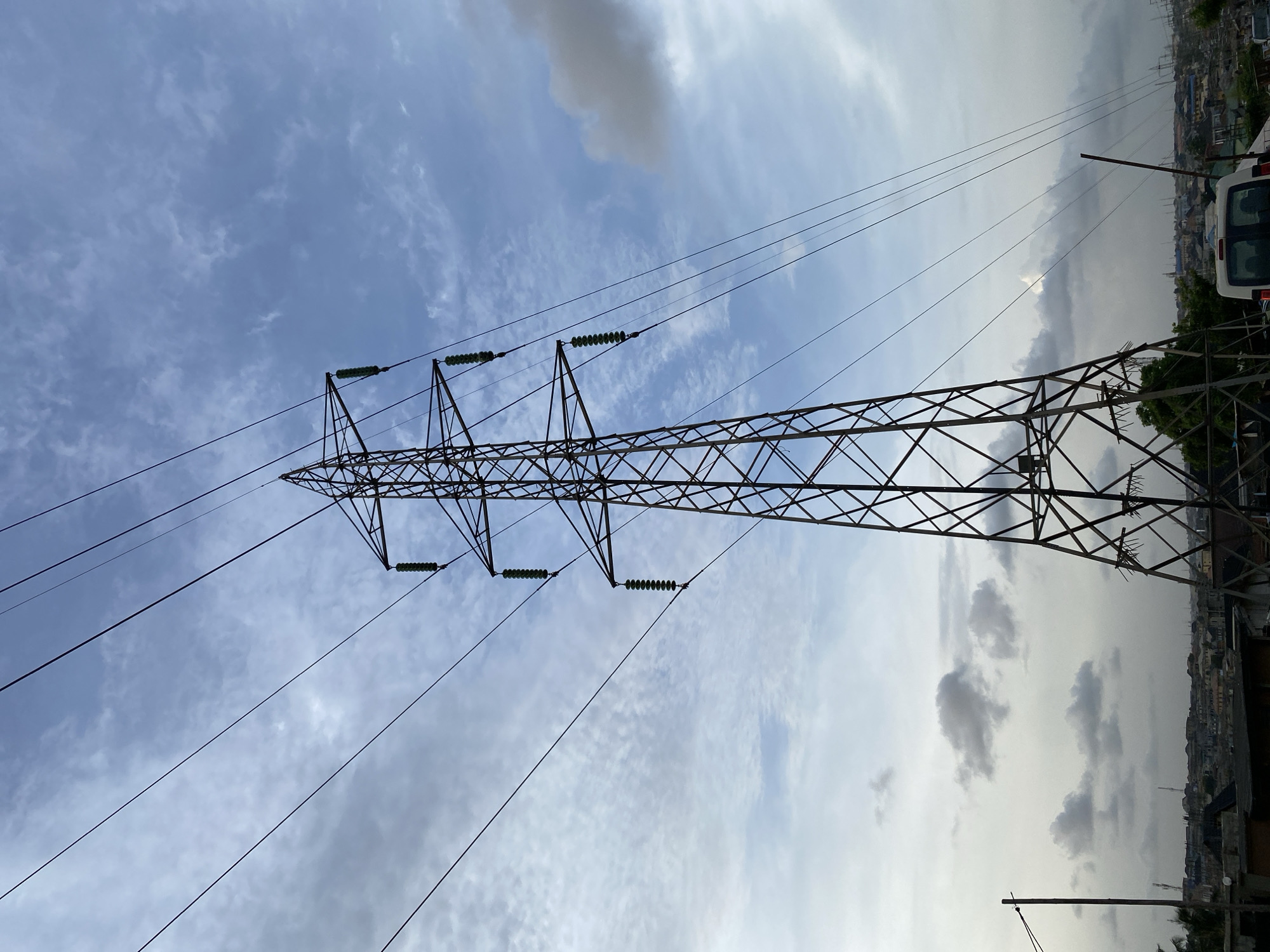 An electrical transmission tower is shown against a partly cloudy sky in an urban environment. The tower is constructed of a dark metal lattice. Attached to the tower are transmission lines, appearing as dark, thin lines extending into the sky. A city can be seen in the background on the right side of the image. A white van is parked in the vicinity of the tower. The sky is mottled with areas of blue and gray clouds. Based on the image context, the location is presumed to be Lagos, Nigeria.