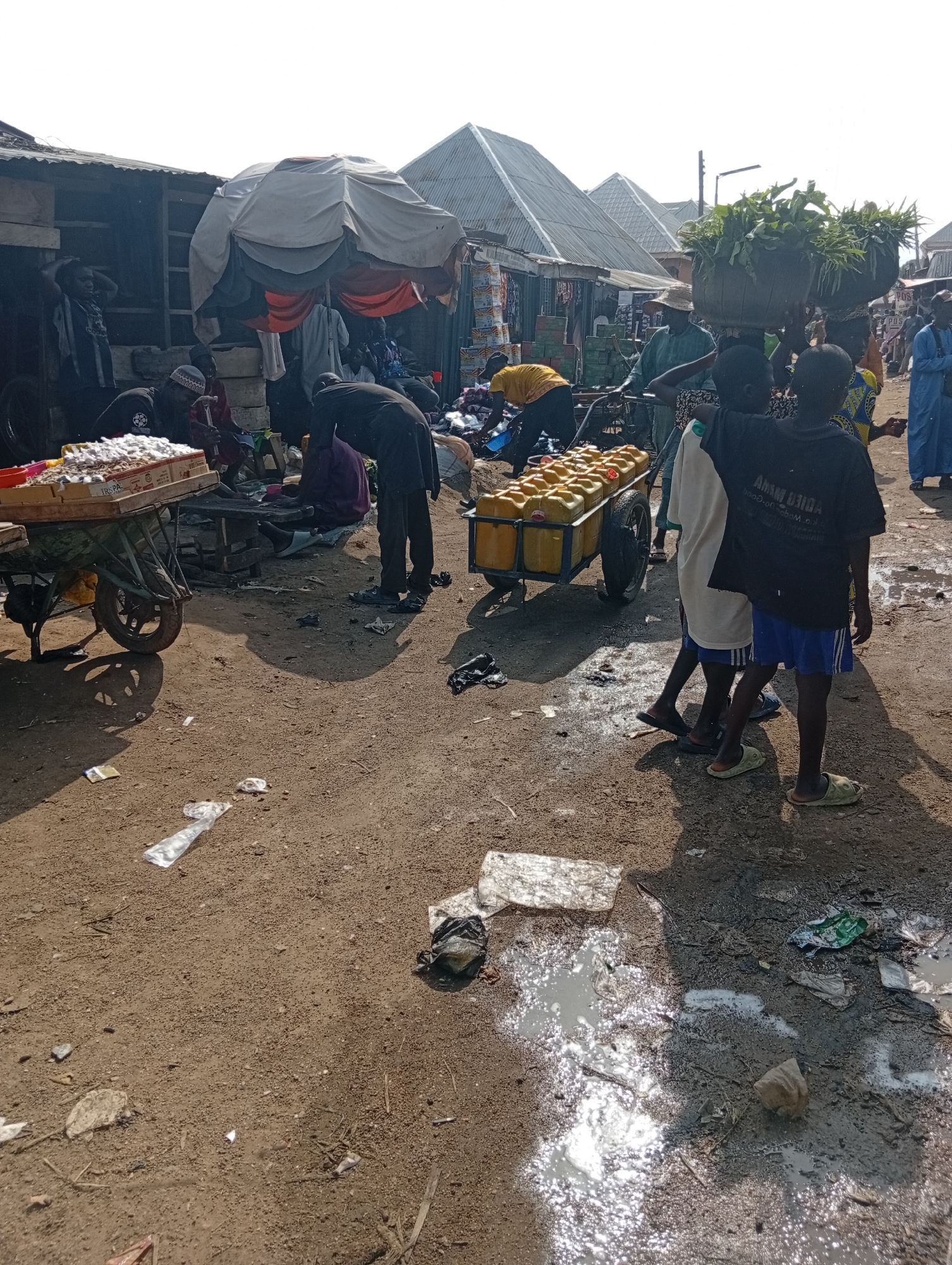 This is a photograph of a bustling marketplace in Jalingo, Nigeria, likely taken during daylight hours. The scene depicts a lively street with several people engaged in commerce and daily activities. In the foreground, a cart is laden with numerous yellow jerrycans, suggesting the sale of liquids such as water or fuel. Beside it, a man in a black t-shirt and blue shorts stands with another person, both looking towards the cart.
Further back, various market stalls and structures are visible, constructed from corrugated iron and wood, typical of informal markets. People are seen tending to their goods, interacting with customers, and moving about. Some individuals are carrying items, including baskets of produce on their heads, indicating a variety of goods being sold. The ground is a mixture of dirt and patchy puddles, with scattered debris, typical of an outdoor market environment. The overall impression is one of a vibrant, somewhat chaotic, but active marketplace.