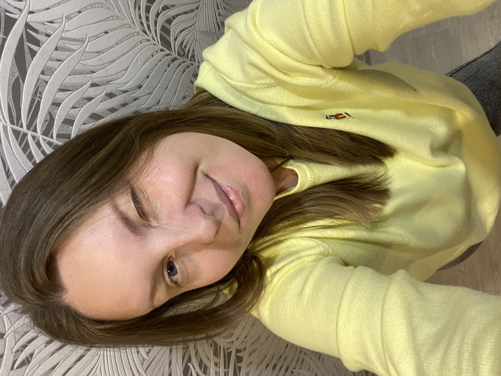 A woman with light brown hair and fair skin is lying down, wearing a yellow sweater with a small emblem on the chest. She is looking at the camera with one eye open and the other closed, suggesting a wink. The background features a patterned surface, possibly wallpaper or a sofa. Part of a greyish material, possibly pants, is visible on the upper right side. The image appears to be a selfie taken indoors. It is not possible to determine if the picture was taken in Noviy, Russia.