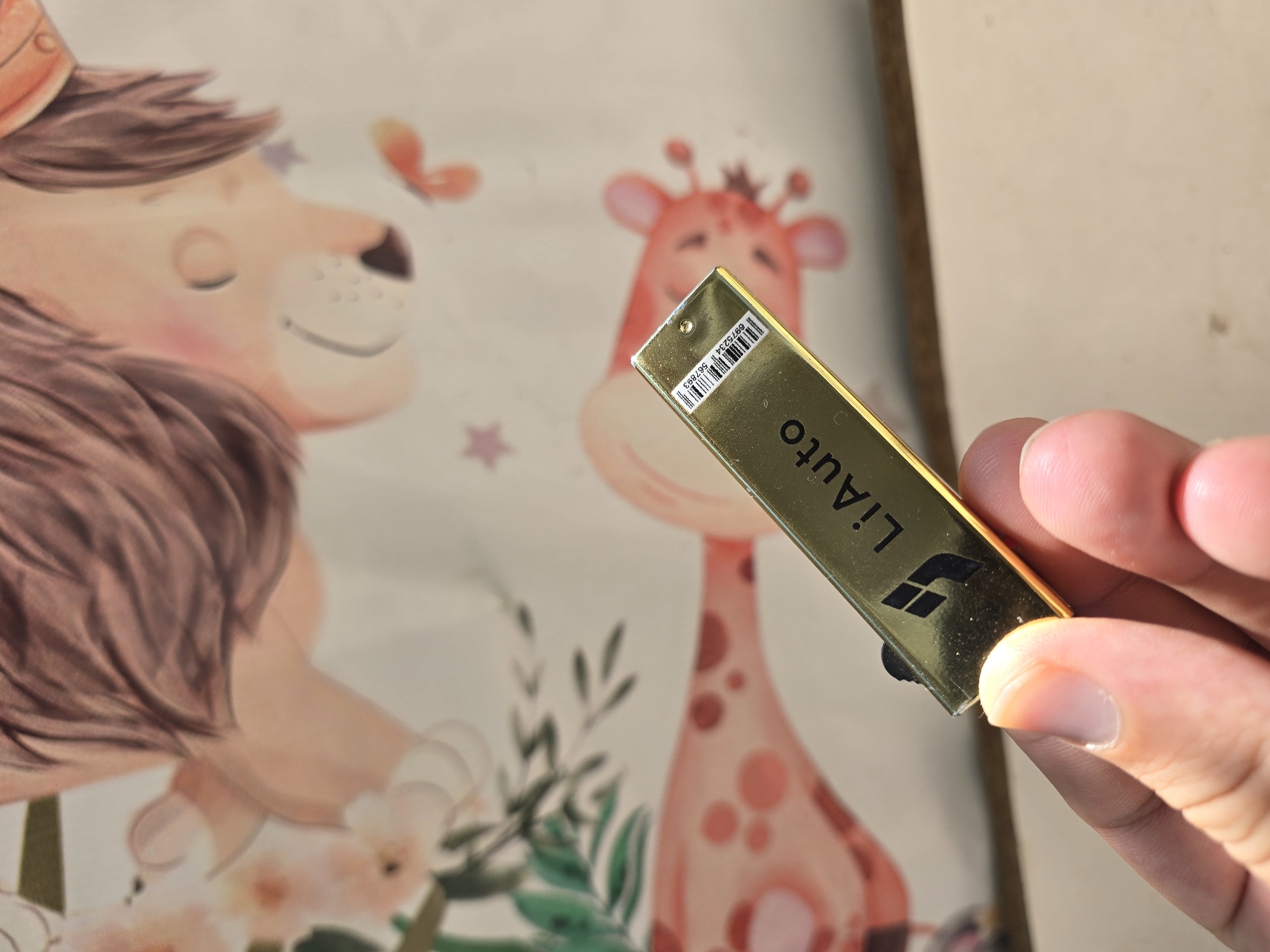 A hand holds a golden USB drive with the brand "LiAuto" visible. The USB drive has a barcode and product number printed on it. In the background, a child's drawing depicts a lion and a giraffe. The image appears to be taken in Qaratau, Kazakhstan.