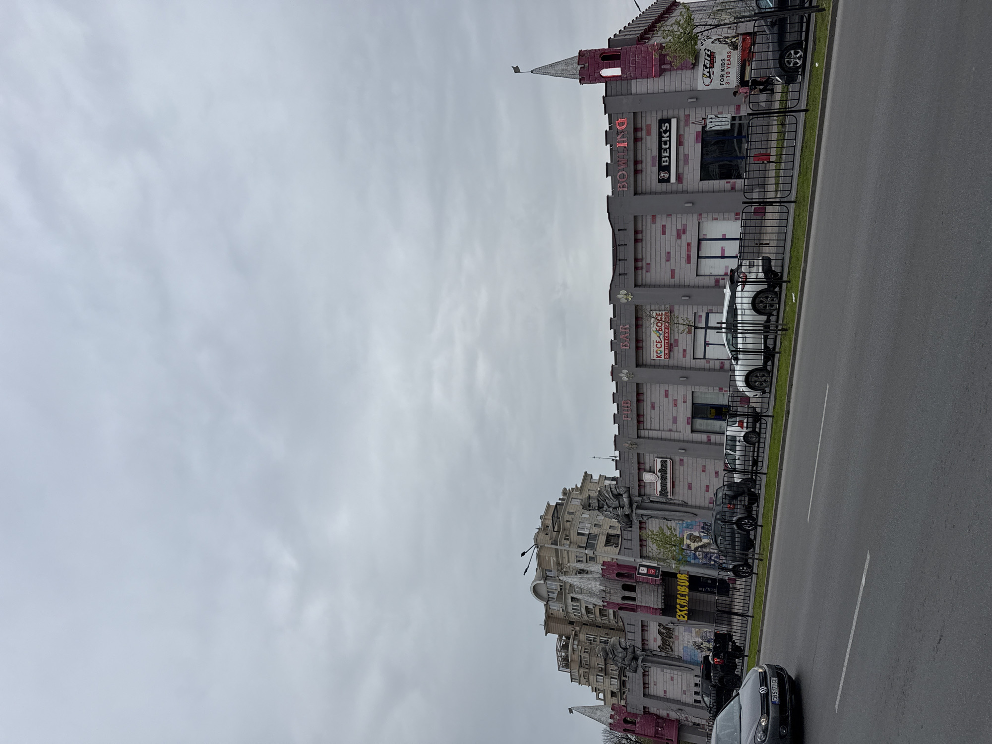 The image captures a street scene in Plovdiv, Bulgaria, focusing on a distinctive building complex designed to resemble a medieval castle. The "castle" houses businesses including a bar, bowling alley ("BECK'S" bowling), and an area for children (kart zone for kids 3-10 years). The building's facade is adorned with grey and red brickwork and castellated features. Signage is prominently displayed.
Parked cars line the street adjacent to the building, partially obscuring the view of its lower sections. A black fence runs along the property's edge. The overcast sky creates a muted, somewhat drab atmosphere. A street runs parallel to the building, complete with road markings. A car with the license plate "CB 5171 CE" is visible on the road. The word "EXCALIBUR" is written in yellow and large letters across the front of the building. Statues of knights stand guard at the castle's entrance.