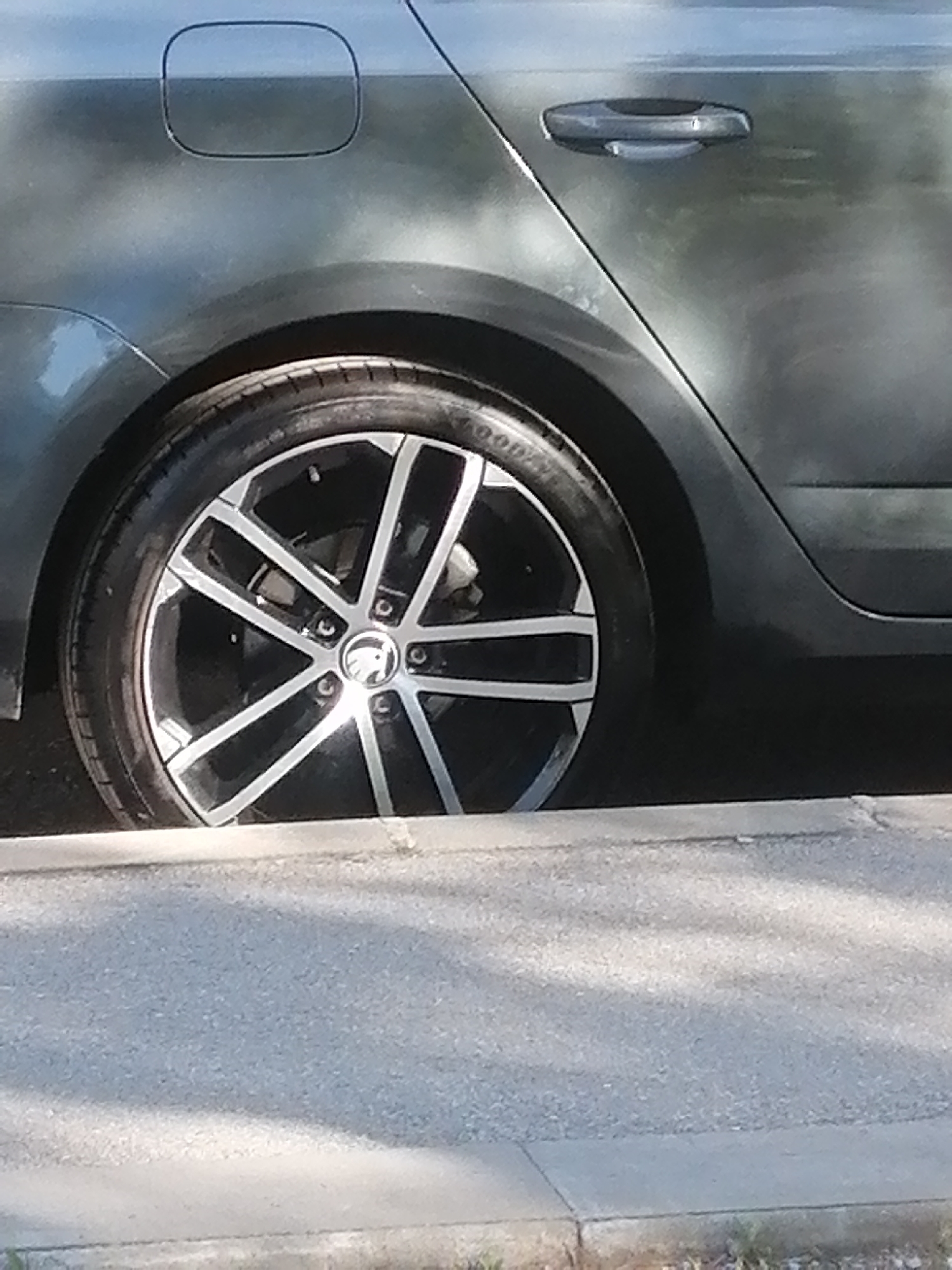 The image shows the rear passenger side of a dark-colored Skoda Octavia parked on a city street. The car is parked parallel to the curb, and the rear tire is touching the curb. The wheel has a multi-spoke design with a Skoda logo in the center. The tire brand is "Goodyear". The car has a fuel filler door visible on the rear quarter panel and a door handle on the rear passenger door. The scene is likely in Sofia, Bulgaria, based on the car model's popularity in Eastern Europe and the style of curb, but there are no definitively Bulgarian elements visible to confirm the location.