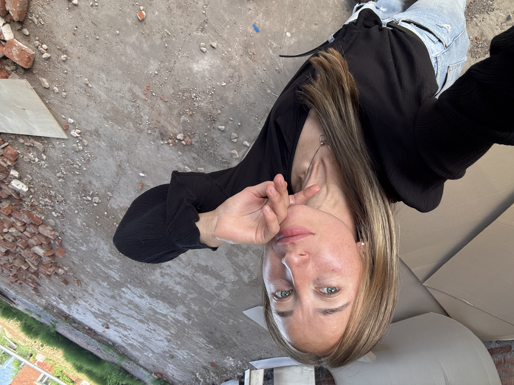 An outdoor, slightly high-angle, full-body selfie of a Caucasian woman with blonde hair is taken at a construction site in Chaikovsky, Russia. The woman is in the foreground, facing the camera and resting her chin on her hand. She is wearing a black, long-sleeved, ruched top and light-wash, ripped denim jeans. Her eyes are blue-green, and her lips are slightly parted. She is wearing a small, square pendant necklace. The background is a rough, concrete ground with scattered bricks and large pieces of cardboard. Green grass and a building with a pipe are visible in the upper right corner. The lighting suggests it is daytime.