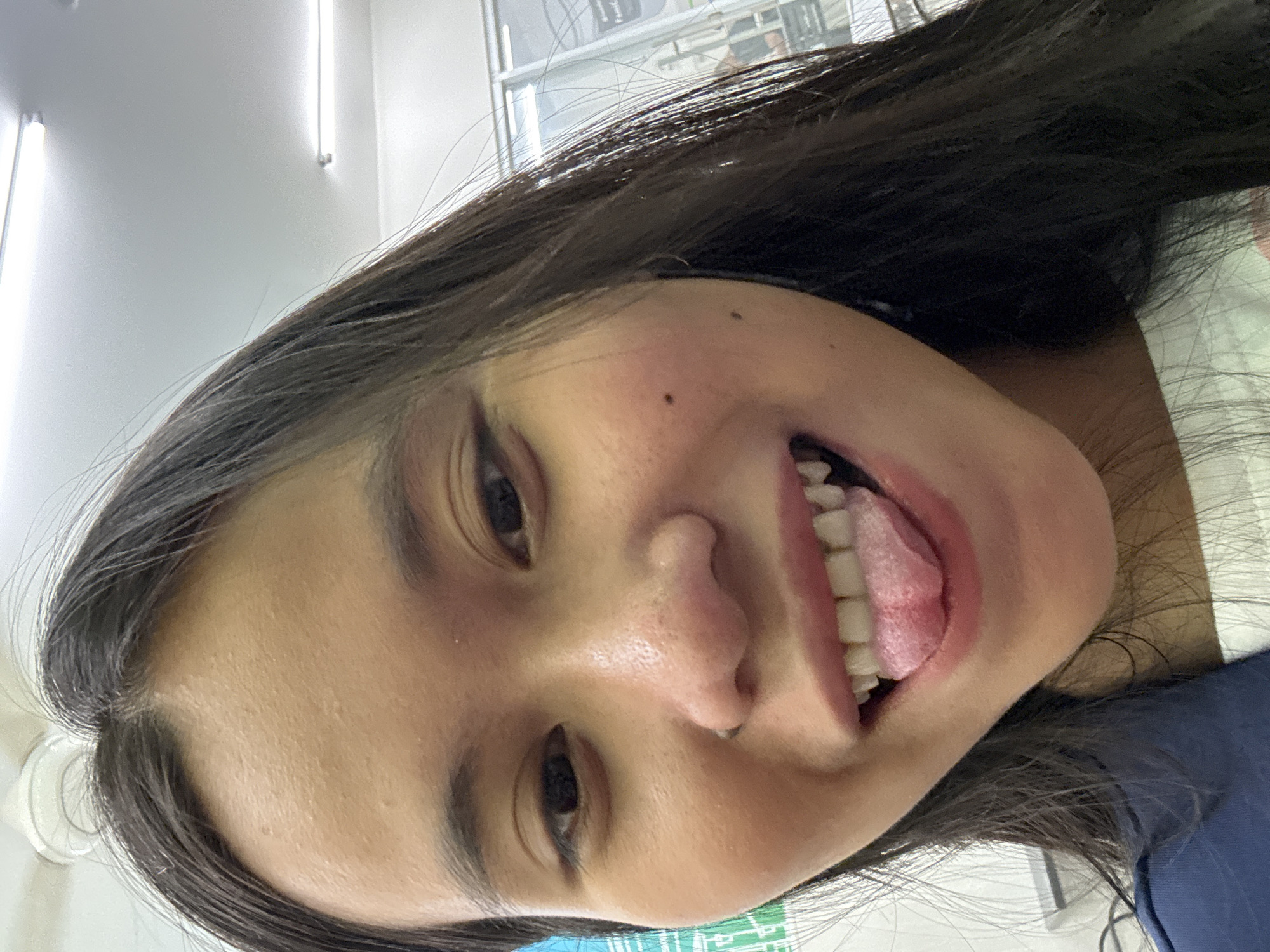 A close-up selfie shows the face of a young East Asian woman with dark hair and brown eyes, sticking out her pink tongue, revealing her white teeth. The woman is wearing a dark blue jacket over a white shirt. Her face is illuminated by overhead fluorescent lights. The background is blurred, but appears to be an indoor setting with fluorescent lights on the ceiling and a glimpse of a store or office with glass walls. The woman has a small nose ring. Her skin has some texture, with visible pores and a few small blemishes. The image quality is clear, with good lighting and focus on the subject. The overall impression is a casual, candid photograph.