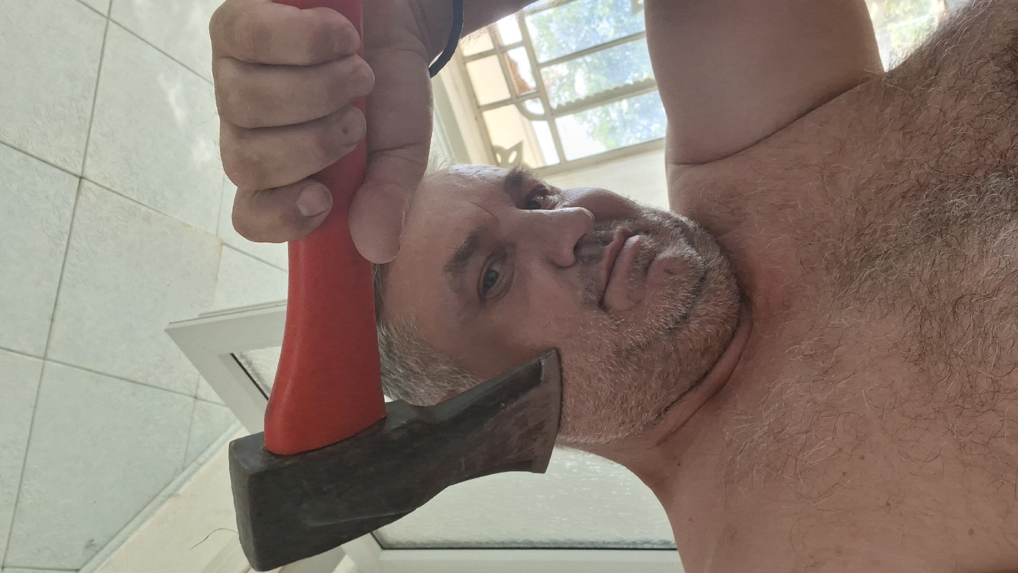 The image is a selfie taken from a low angle, showing a man holding a red-handled axe near his head. The man appears to be middle-aged with graying hair and a beard. He is shirtless and his chest is hairy. The background shows a tiled ceiling and a window with greenery visible outside. The location is specified as Voinova, Moldova. The man is looking directly at the camera with a neutral expression. The axe is positioned so that the blade is close to his left temple.