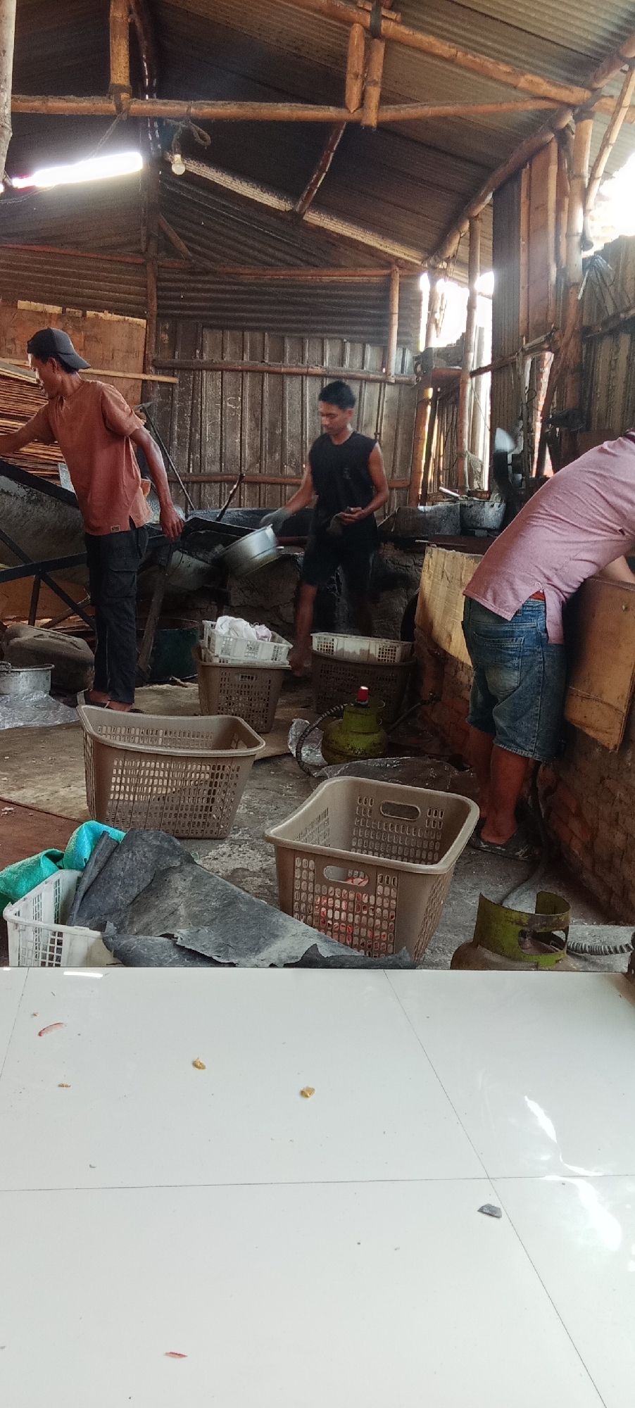 The image depicts an indoor scene, likely a workshop or processing area, in Kesamben, Indonesia. The environment is characterized by a rustic, open-air structure with a corrugated metal roof supported by bamboo poles and wooden beams. The walls appear to be made of corrugated metal and wood. The floor is concrete, with some areas appearing wet or dusty.
Three individuals are present in the scene. In the foreground, to the left, a man wearing a reddish-brown t-shirt and dark pants is seen from the side, facing a large metal basin or vat. He is wearing a dark baseball cap and appears to be engaged in some form of manual labor.
In the middle ground, a second man, wearing a black sleeveless t-shirt and dark shorts, is observed from the side, walking and holding a metallic ladle or scoop. He is wearing gloves. He is near another large metal vat.
In the right side of the image, a third person, whose face is not visible, is bending over a large wooden structure. This individual is wearing a pinkish t-shirt and blue denim shorts, and also appears to be engaged in work.
Various objects are scattered around the space, including several large plastic baskets of different colors (brown, white) containing what appears to be processed material or raw ingredients. Two green gas cylinders are also visible. A large piece of dark, flexible material, possibly plastic sheeting or tarp, is draped over a white basket in the foreground. The overall impression is one of a functional workspace involved in some kind of production or processing activity.