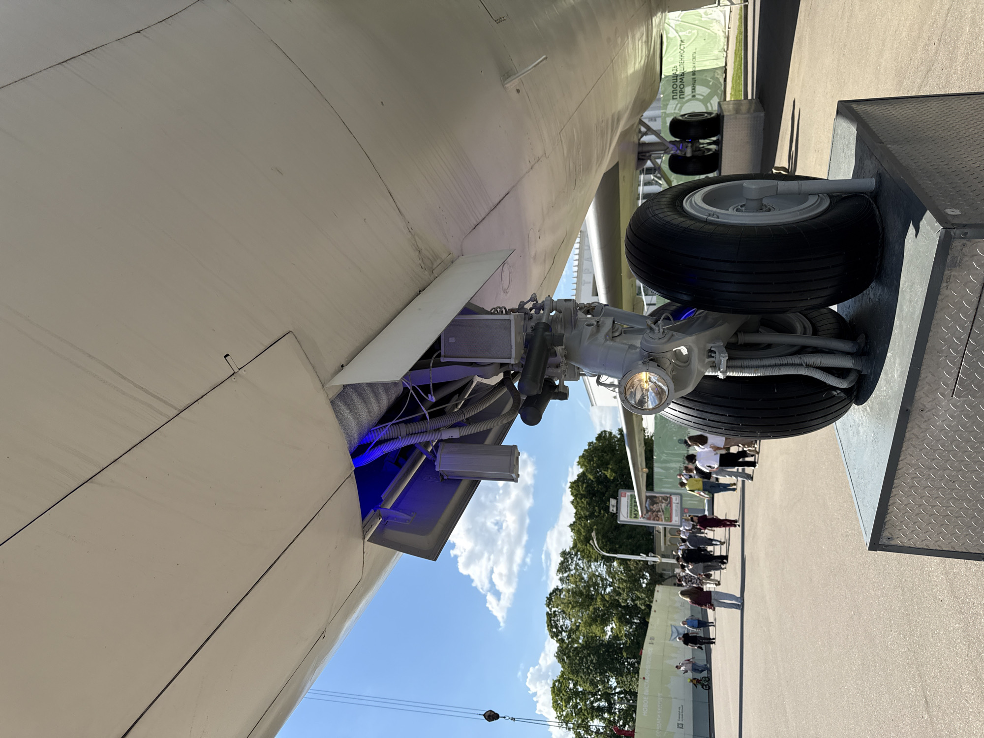An outdoor scene captured from a low angle shows the underside of a large, light-colored aircraft, likely an airliner or cargo plane, displayed in Khimki, Russia. The main landing gear, featuring a black rubber tire on a gray rim and a prominent landing light, is fully extended and rests on a textured, metallic platform. The aircraft's open landing gear bay reveals a complex network of wires, hoses, and mechanical components, illuminated by a distinct blue light emanating from within. A rectangular grey box is mounted inside the bay.
In the background, under a bright blue sky with scattered white clouds, numerous people are visible, standing and walking on a paved surface, seemingly observing the aircraft. Large, green-leafed trees frame the left side of the background, and various exhibition structures or buildings are partially visible. One green banner in the distance displays Cyrillic text, including "ПЛОЩАДЬ ПРОМЫШЛЕННОСТИ" (Industrial Square/Area) and "НОВОЕ В ВДНХ. ЭКСПОЗИЦИИ" (New at VDNKh. Exhibitions), indicating a connection to the well-known VDNKh exhibition center.
The scene suggests an aerospace exhibition or museum display on a clear, sunny day, likely mid-day or afternoon. The aircraft's fuselage exhibits some dirt streaks and wear, common for outdoor static displays. A crane hook and cable are partially visible in the bottom left, suggesting the aircraft's installation or setup. The image conveys a calm, observational atmosphere, focusing on the engineering details of the aircraft's landing gear.