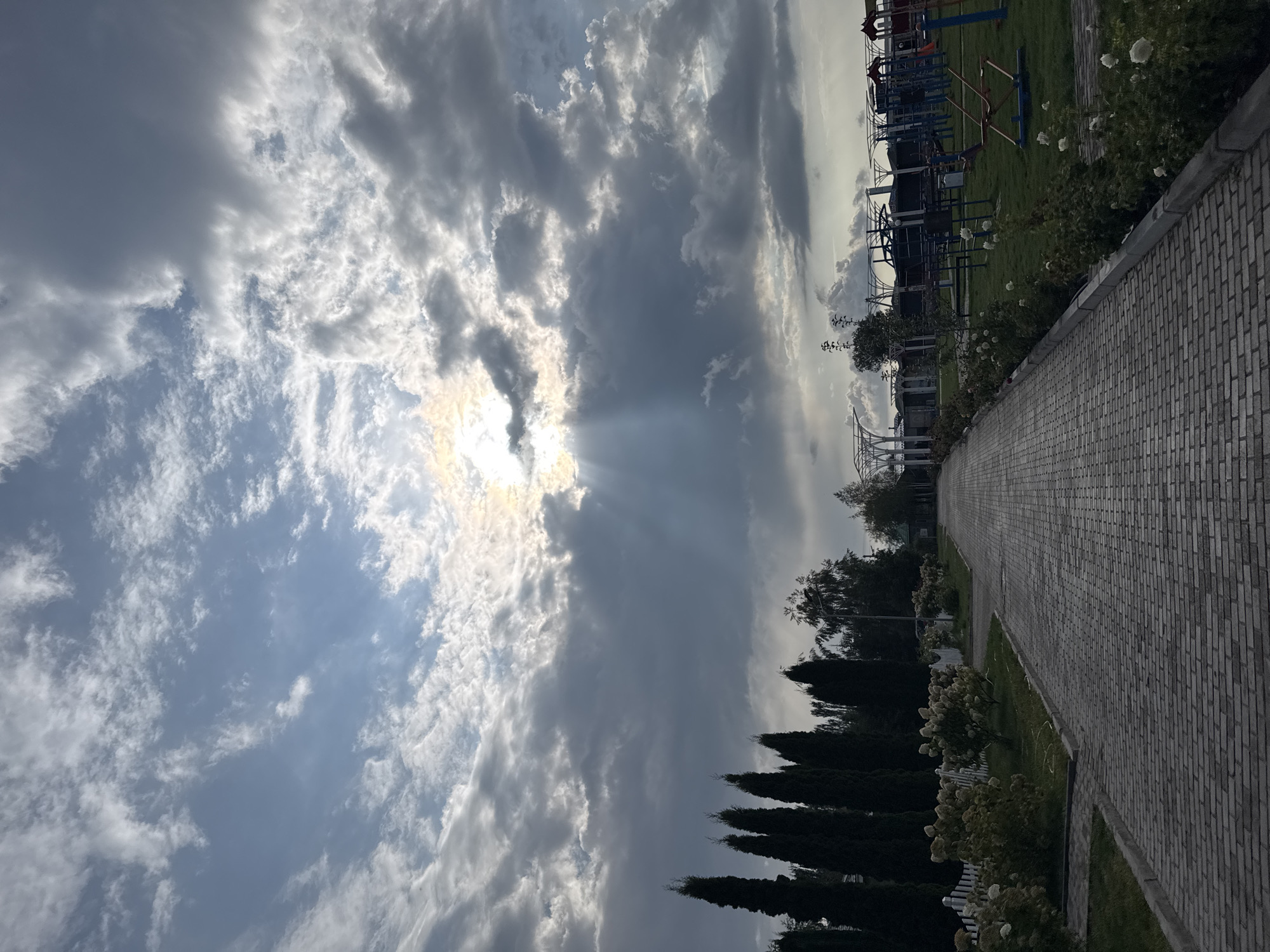 The image depicts a paved pathway leading into what appears to be a park or recreational area in Sary-Oy, Kyrgyzstan. Tall, dark green cypress trees are visible on the left side of the pathway, bordering a white picket fence and flowering bushes. The pathway itself is constructed of light gray bricks. On the right side, the pathway borders a grassy area with a playground visible in the background, featuring colorful structures like swings, slides, and climbing frames. The sky above is dramatic, with a mixture of bright sunlight breaking through scattered clouds, casting rays of light downwards. The overall lighting suggests it may be late afternoon or early evening. No people are visible in the scene.
