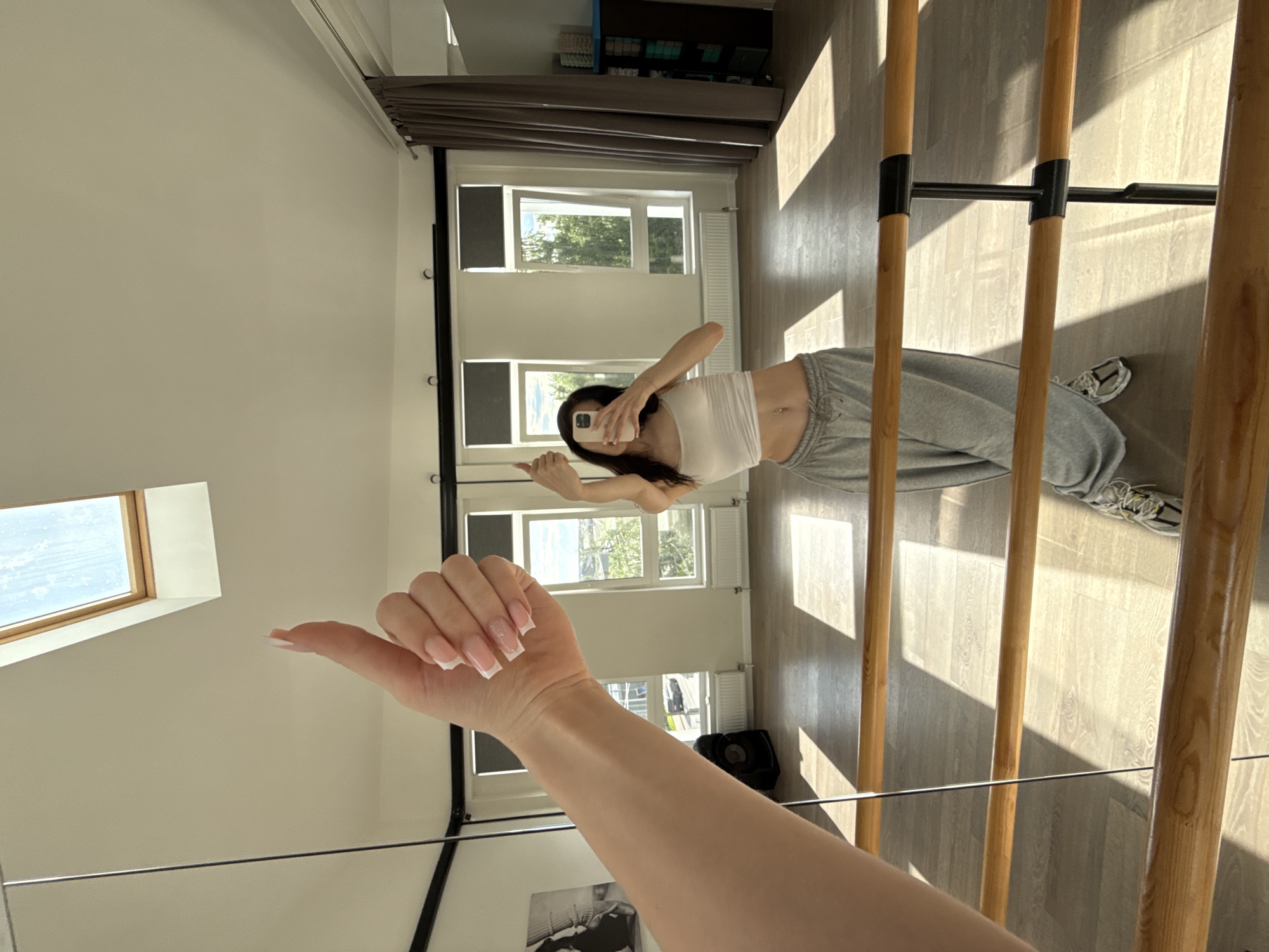 The image is a mirror reflection of a woman in a dance studio in Zakharovo, Russia. The woman is wearing a white crop top and grey sweatpants, and is holding her phone to take a selfie. She has long, dark hair and a slim build. Her nails are long and painted with a French manicure. In the foreground, her right arm is extended towards the camera, with her thumb up.
The dance studio has wooden floors and a barre, typical of a ballet or dance studio. There are large windows that let in natural light, suggesting it is daytime. The shadows cast by the barre and window frames indicate a sunny day. Through the windows, trees and greenery are visible, suggesting a location surrounded by nature. In the reflection, the room appears to be well-lit and spacious. The overall mood is casual and relaxed, with the woman appearing confident and at ease. There is no visible text in the image.