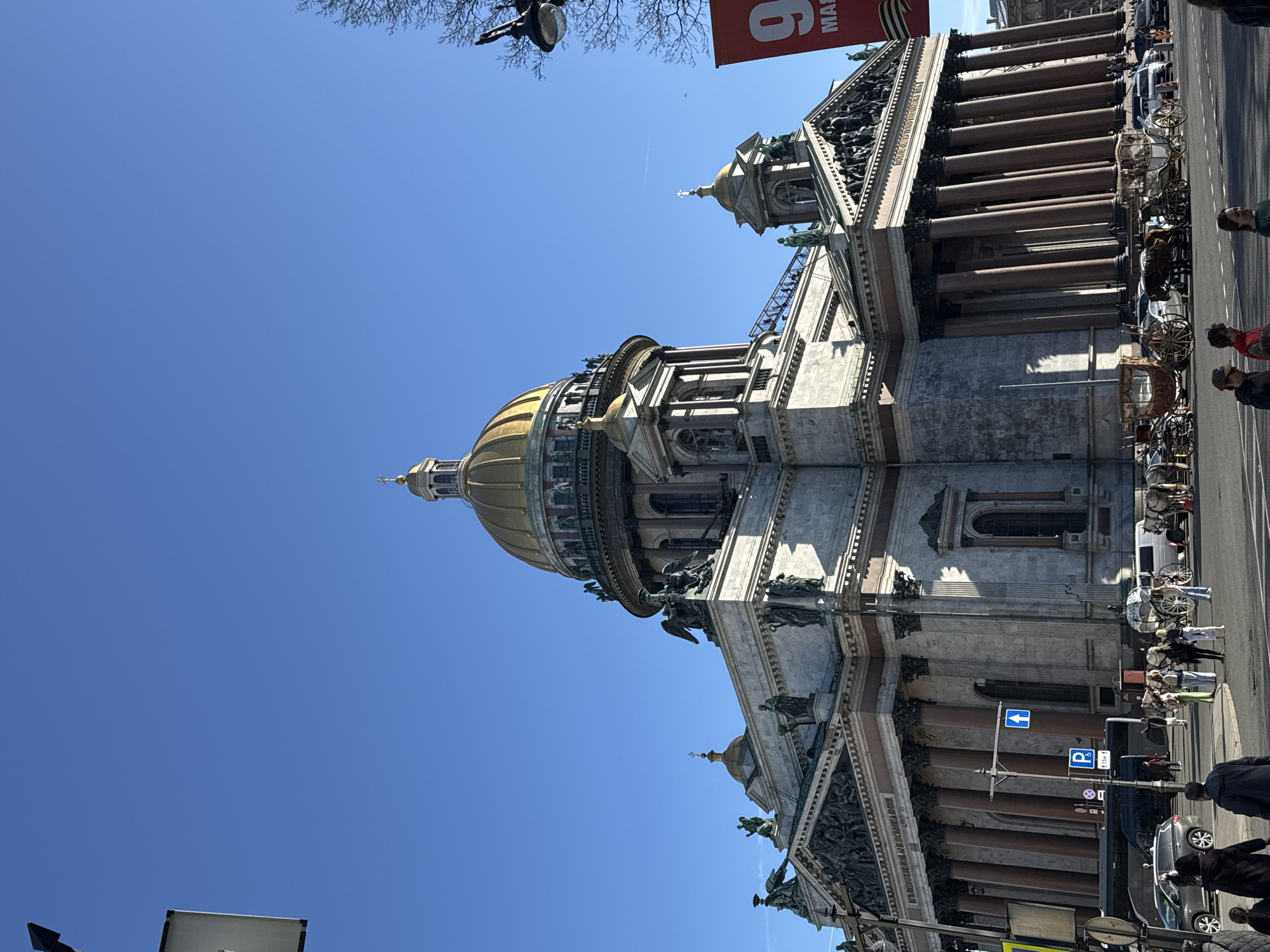 This image captures a vibrant street scene in Saint Petersburg, Russia, dominated by the majestic St. Isaac's Cathedral under a clear, bright blue sky. The grand cathedral, with its prominent golden dome and several smaller cupolas, features classical architecture, including large columns and intricate sculptures on its facade.
On the street level to the right of the cathedral, several horse-drawn carriages, some with horses and some empty, are visible, suggesting tourist activity. People are present on the pavement, some near the carriages, others walking. Parked cars are also seen further down the street. The scene appears calm and bustling, indicative of a regular day.
The weather is sunny with no clouds, suggesting a pleasant daytime. A red banner is partially visible in the upper right, displaying the number "6" and the word "МАЯ" (May), along with a St. George's ribbon, a symbol often associated with Victory Day (May 9th) in Russia. This detail helps to place the timeframe of the photo around early May. Street signs include a blue arrow pointing left and a blue 'P' parking sign with "15 min" underneath. The overall atmosphere is lively, showcasing a prominent architectural landmark and local street life.