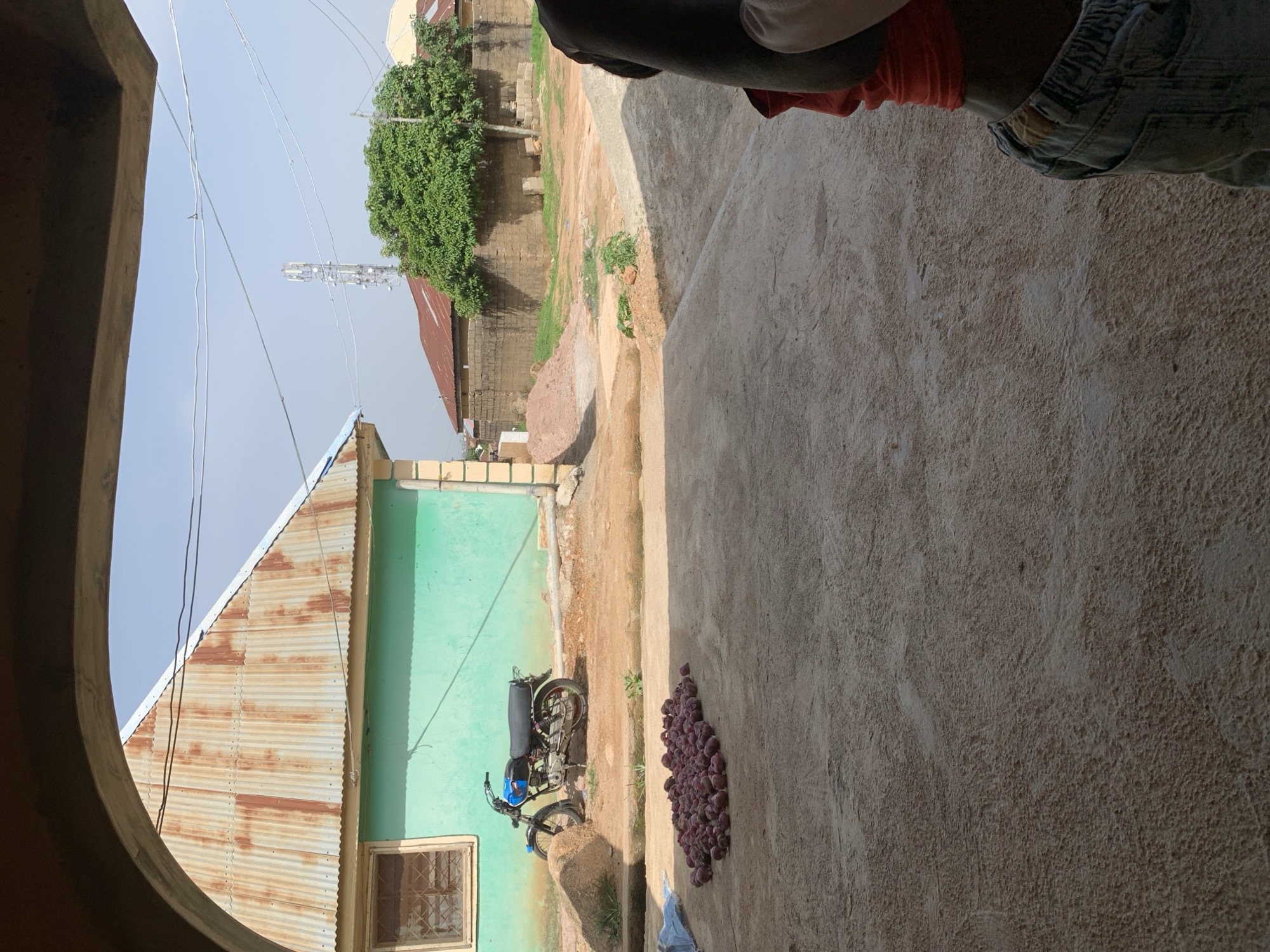 An outdoor scene in the city of Jos, Nigeria, shows a collection of red onions spread out on a dirt ground. To the left of the onions, a motorcycle is parked near a teal-colored building with a corrugated metal roof. In the background, a cell phone tower is visible, along with other buildings and trees. The foreground is dominated by a concrete surface. A person, only their torso and lower body visible, is standing on the right side of the frame, facing away from the camera. The person is wearing a red shirt and blue jeans. The sky is clear and blue.