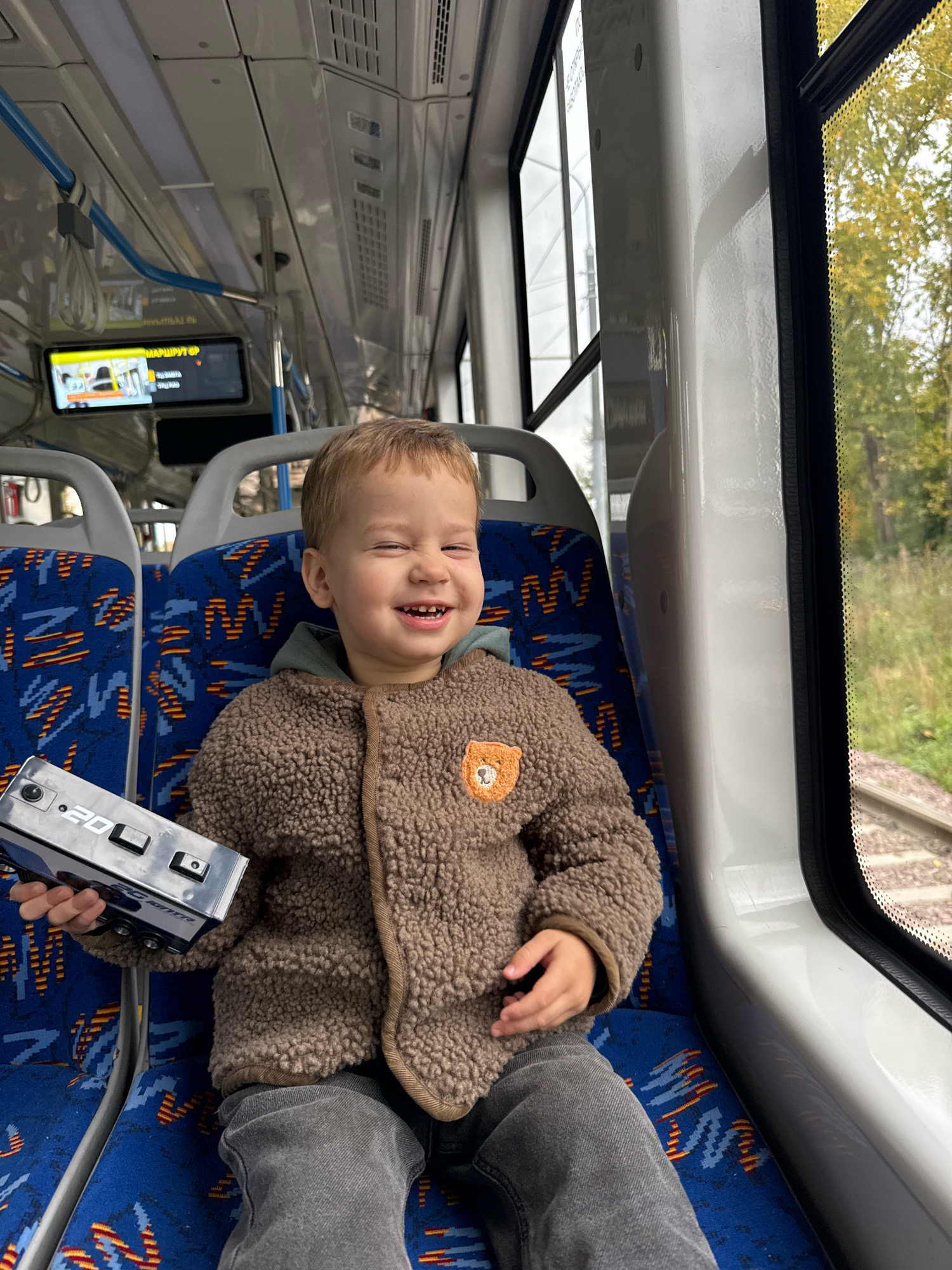 A young boy is seated on a blue bus seat with a patterned design, holding a silver object with buttons and the number "20" on it. He is wearing a brown, fuzzy jacket with a teddy bear patch on the chest and grey pants. The boy is smiling with his eyes closed, looking to his right. The interior of the bus shows grey walls, blue poles, and a digital screen displaying route information. Outside the window, trees and train tracks are visible. The image appears to be taken from inside the bus, looking towards the boy and the window.