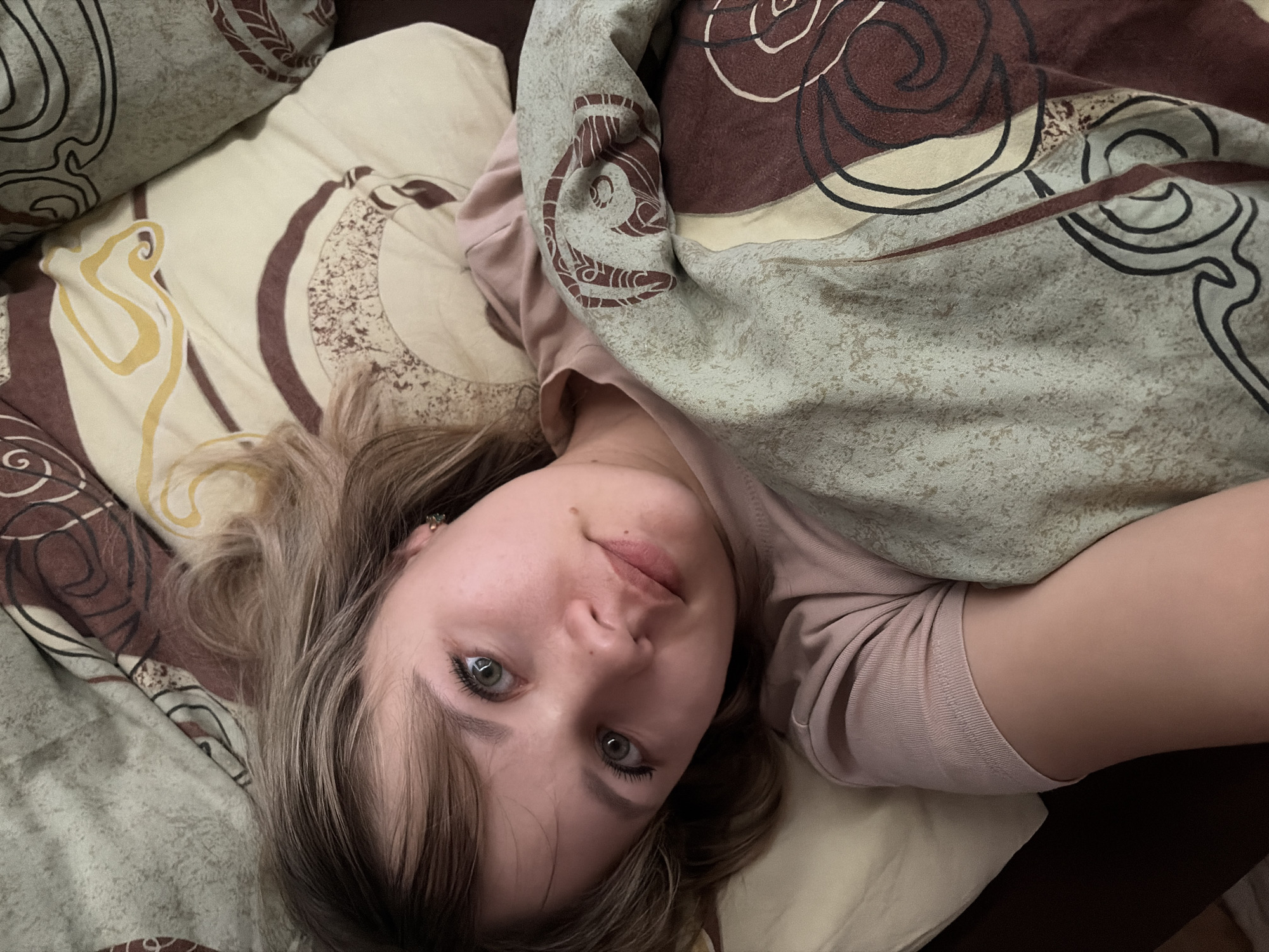 An overhead shot captures a young person, appearing to be female, lying in bed, looking directly up at the camera. Her head is positioned on a pillow towards the upper left of the frame, with her body extending downwards and to the right, mostly covered by bedding.
She has light brown to blonde hair, parted roughly in the middle, falling around her face and on the pillow. Her eyes are open and appear to be green or hazel, with dark lashes. Her expression is neutral and calm, with a slight hint of a subtle smile. Visible facial features include a few small moles or freckles on her nose and cheek. She is wearing a light pink or mauve long-sleeved top or t-shirt.
She is partially covered by a duvet or blanket that is predominantly light green-grey with a distressed texture, featuring abstract swirly patterns in brown and black lines. The pillow and other bedding visible have a similar design, with swirly, organic patterns in shades of beige, brown, and yellow, with black outlines. The overall impression is that of someone relaxing or waking up in a bedroom. There is no visible text in the image.