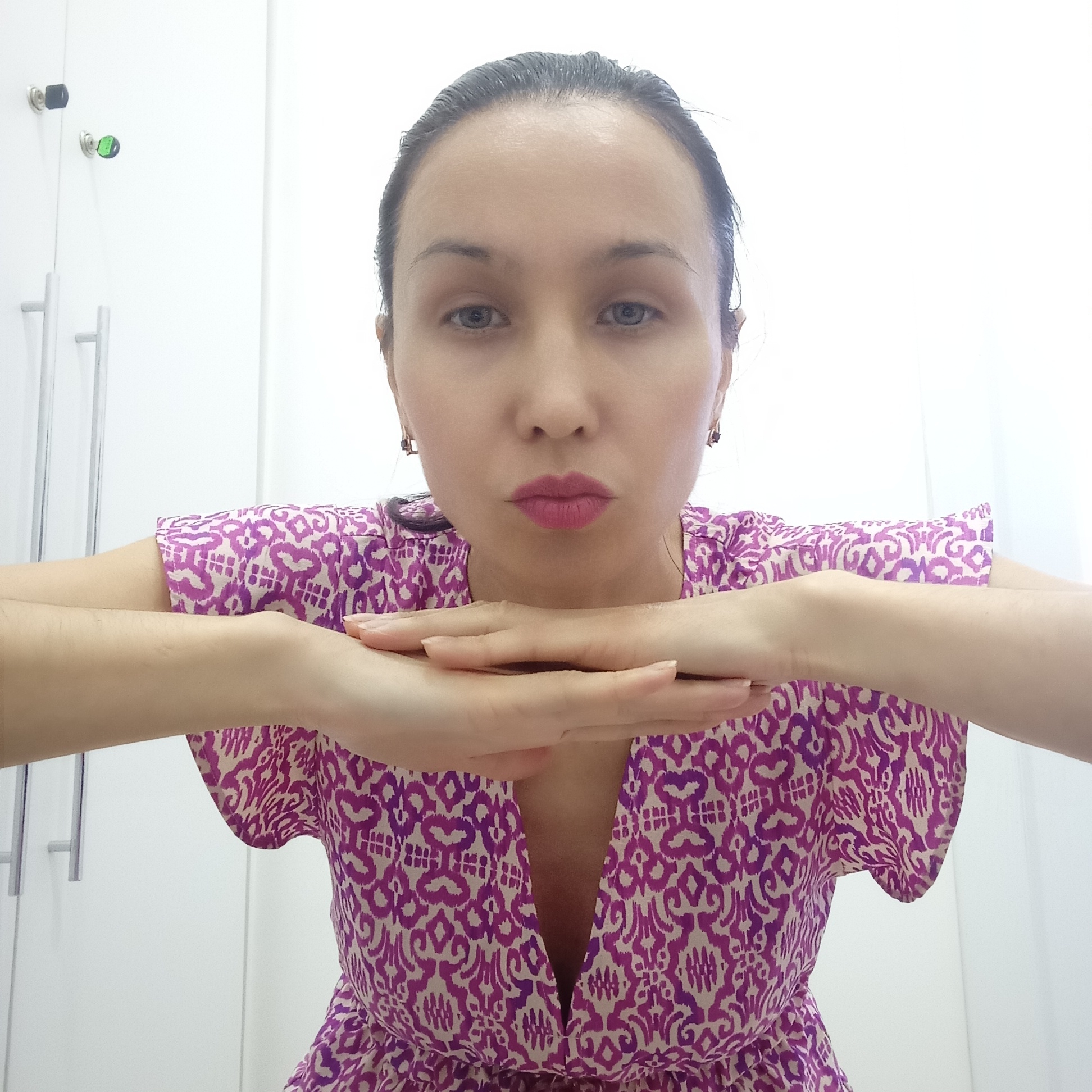 A woman with fair skin, dark hair pulled back, and wearing a patterned purple and white dress is looking directly at the camera. Her hands are crossed in front of her, supporting her chin. The background is a white wall with a cabinet that has silver handles and what appear to be a green and a black key-like object in the upper left corner. There is no information in the image to indicate the location is Izhevsk, Russia.