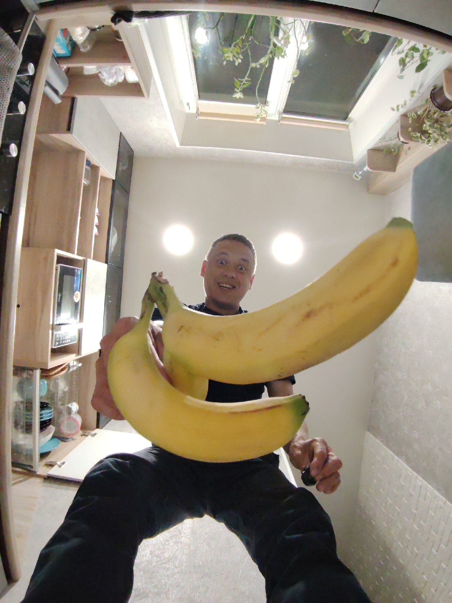 A man holds two large bananas in front of his face, taking a low-angle shot with a fisheye lens. He wears black pants and a black shirt. The background shows a kitchen interior with wooden cabinets, a microwave, and plants visible through a skylight. The location is potentially in Chaikovsky, Russia.