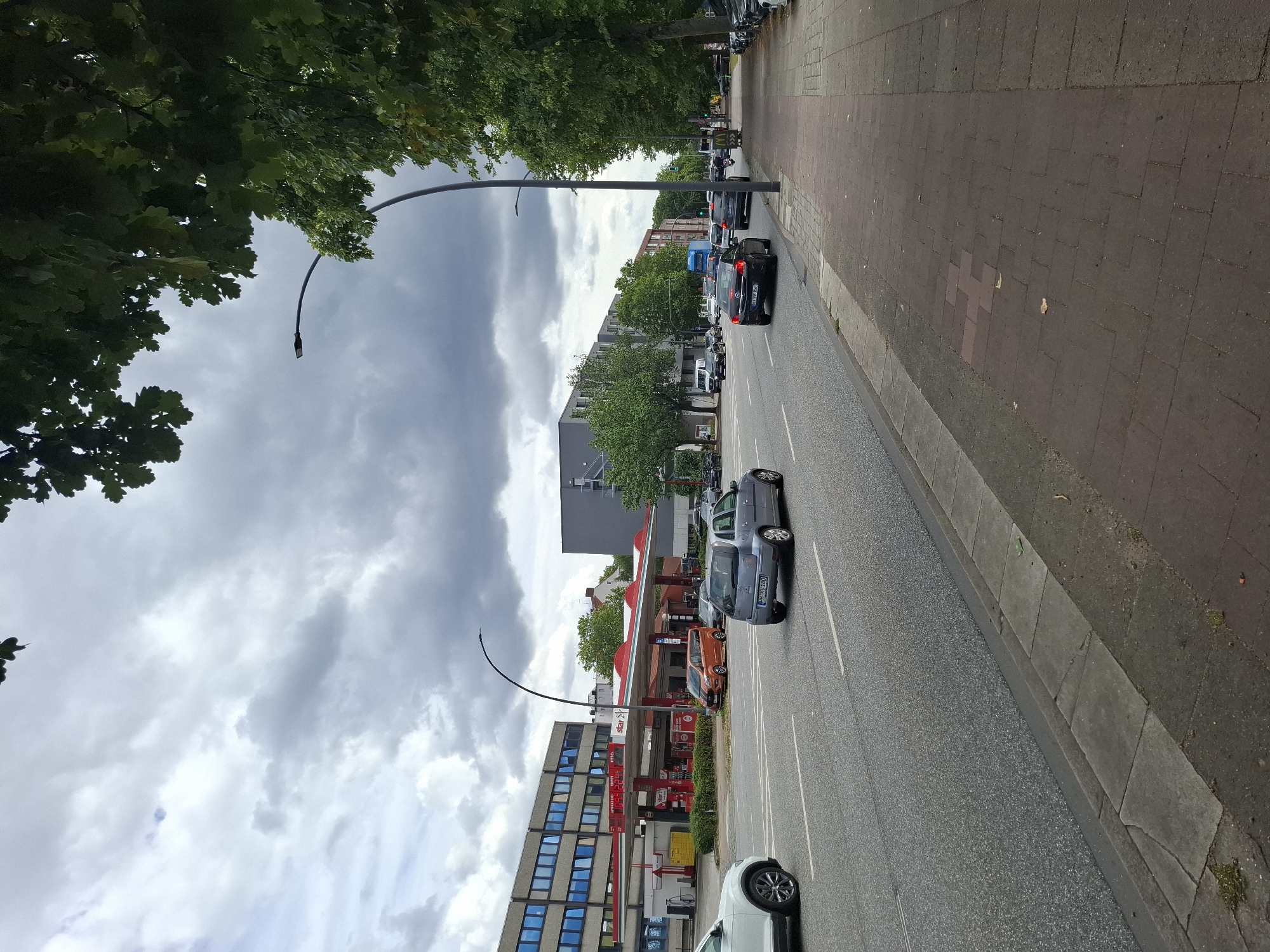The image depicts a street scene in Hamburg, Germany, specifically on a cloudy day. A multi-lane road runs horizontally across the frame, with cars driving in both directions. On the left side of the road, there is a gas station with the "star" logo. Beyond the gas station, there are modern buildings with blue-tinted windows. On the right side of the road, a tree-lined sidewalk is visible with pedestrians and parked cars. The sky is overcast with dramatic clouds. The overall impression is of an urban environment with everyday activity.