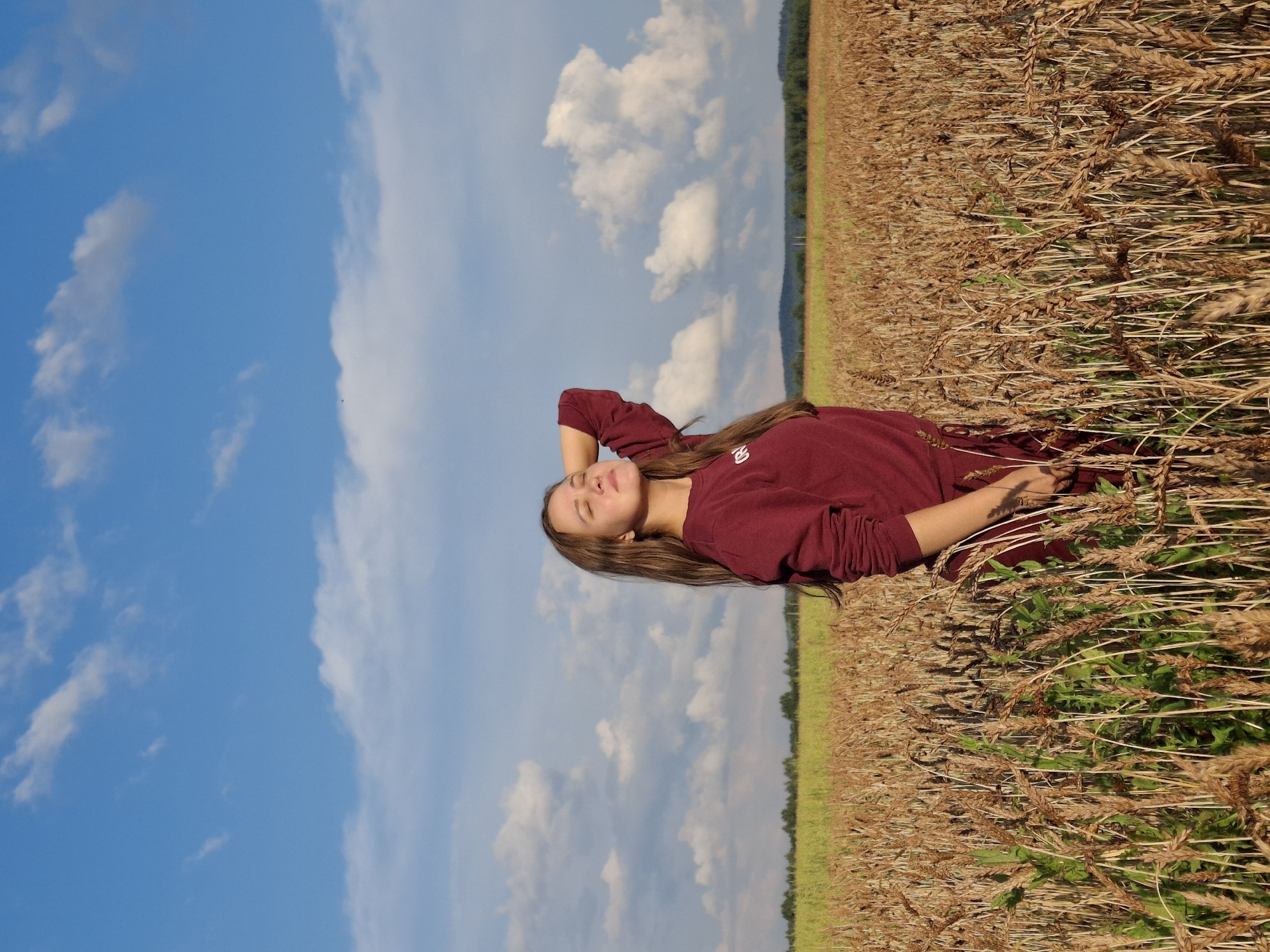 A young woman with long brown hair stands in a field of wheat under a blue sky with white clouds. She is wearing a maroon sweatshirt and has her right arm raised behind her head, with her eyes closed and her face tilted towards the sun. The wheat field extends into the background, with a line of trees visible on the horizon. The image appears to be taken outdoors in a rural setting, possibly in Plishkino, Russia, given the presence of a vast wheat field characteristic of the region.