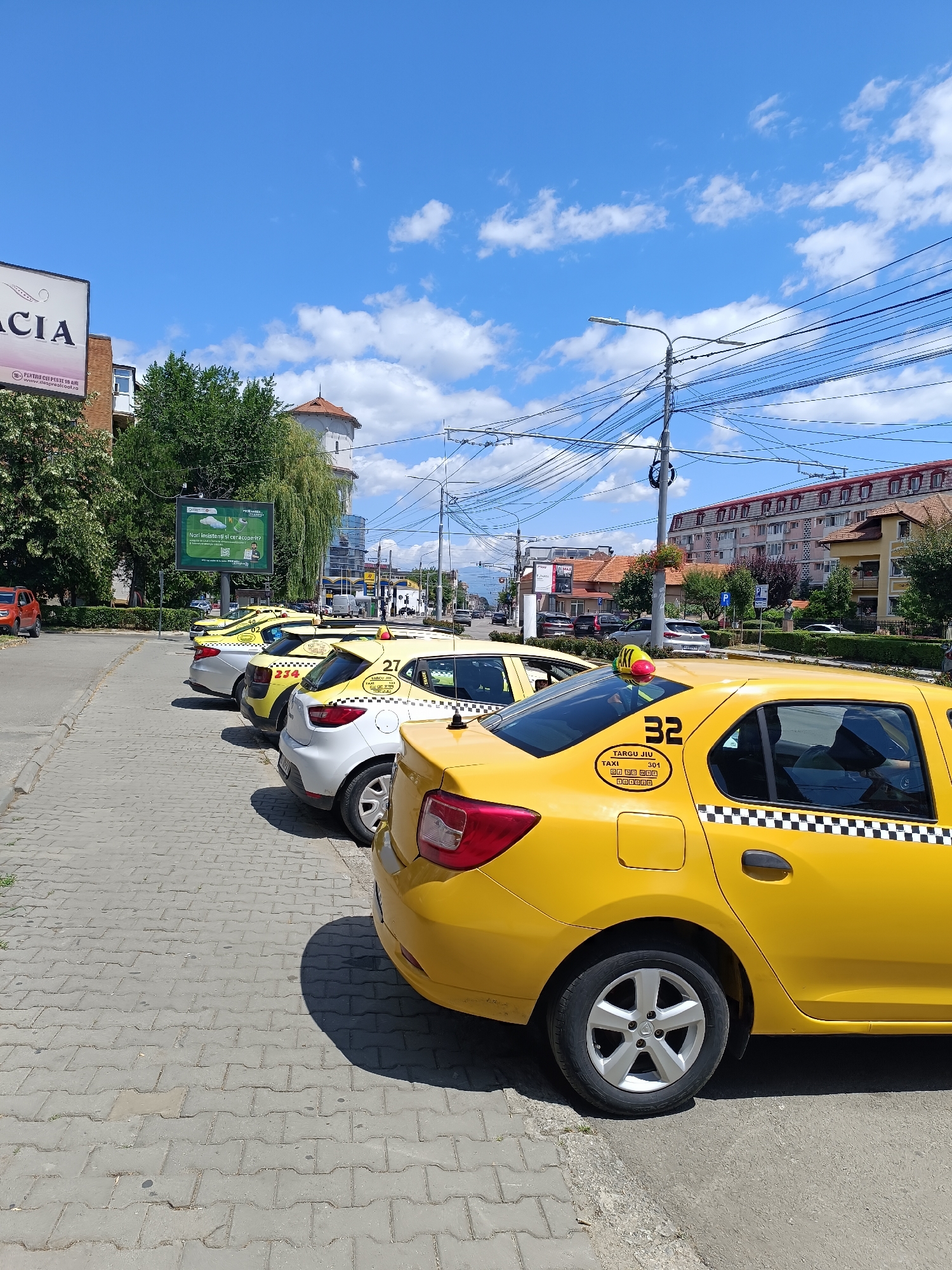 This image shows a row of yellow taxis parked on a street in Târgu Jiu, Romania. The taxis are lined up on a paved sidewalk next to a road with a few cars driving in the background. The sky is blue with some white clouds, and there are trees and buildings visible on either side of the street. The taxi in the foreground has the number "32" on its door and displays "TÂRGU JIU" and "TAXI 301" on a sticker. The other taxis in the row also have numbers and markings, with some of them being partially visible. A large green billboard with advertisements is visible in the background, to the left of the taxis. The overall scene appears to be a typical urban streetscape on a sunny day.
