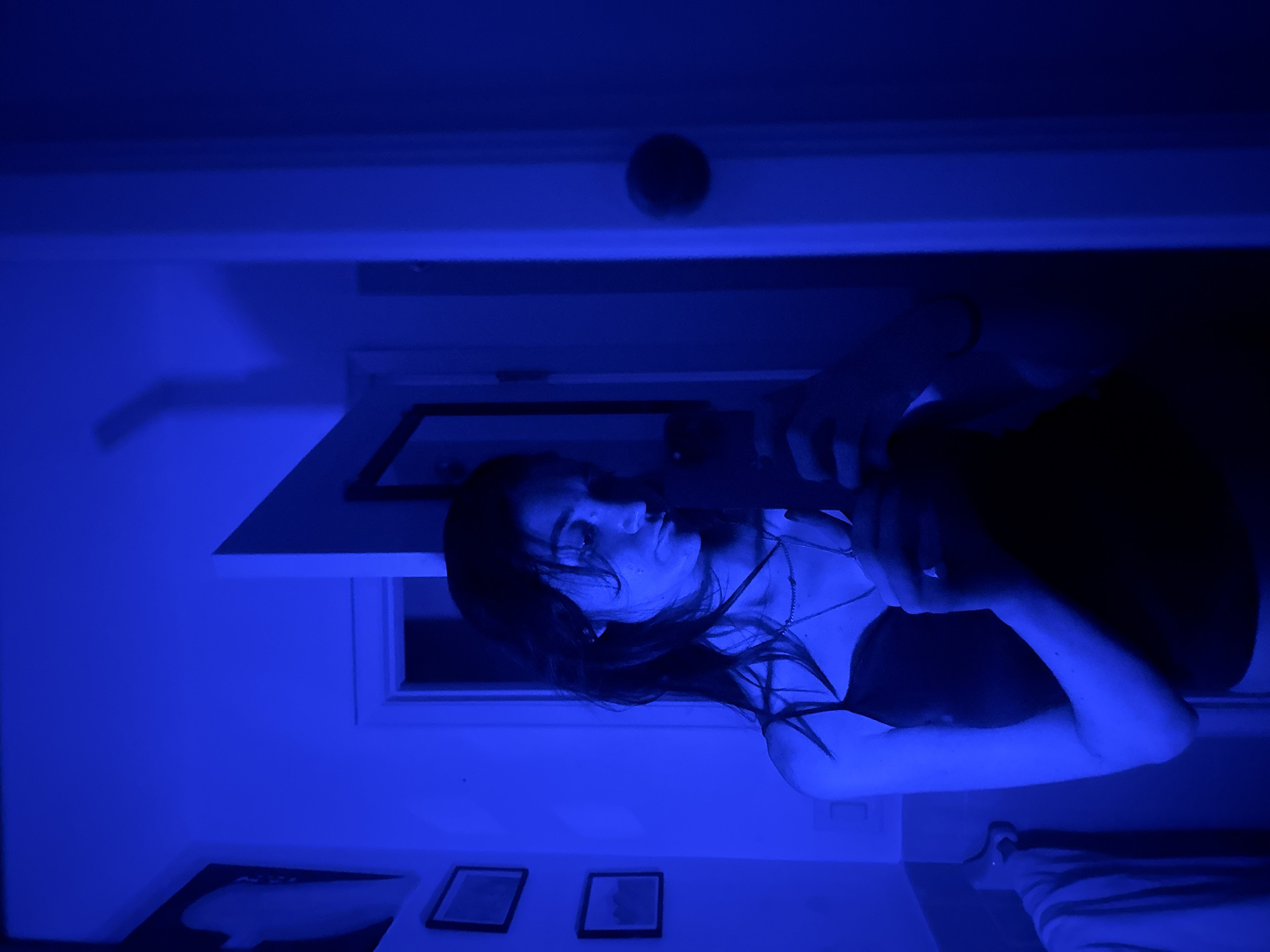 The image captures a person, appearing to be a young woman with long dark hair, indoors under pervasive, deep blue lighting. The entire scene is bathed in a strong, monochromatic blue, creating a cool, moody, and atmospheric ambiance, suggesting it is evening or night and illuminated by artificial blue light sources.
The woman is positioned centrally, looking slightly off-camera with a contemplative or serene expression. She wears a dark-colored top with thin straps and a necklace featuring a small pendant. Her right arm is raised, holding what appears to be a smartphone, suggesting she is either taking a photo or video, possibly a selfie. Her left arm is bent, resting closer to her body.
In the background, a white door frame or a large window frame is visible behind her. Below it, on the wall, are two framed pictures or decorations, with a third, larger one partially visible to the far left. On the ceiling, a circular object, possibly a smoke detector or light fixture, is discernible. To the right, a striped textile, potentially part of a blanket or towel on a bed or sofa, is partially visible. No discernible text is present in the image. The setting is a generic indoor space, located in New York, United States of America.