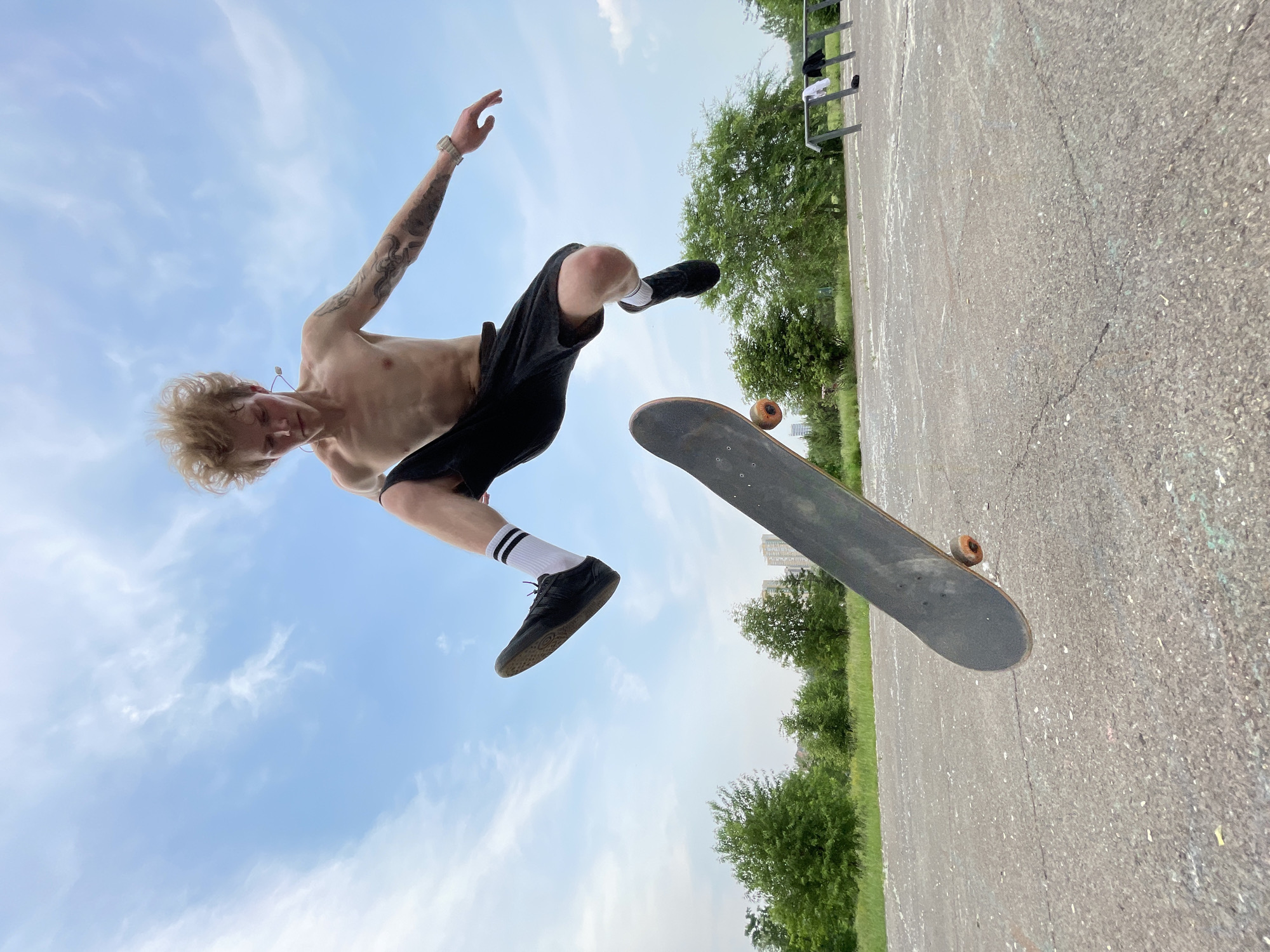 The image shows a young man in mid-air performing a skateboarding trick. He is shirtless, wearing black shorts, white socks with black stripes, and black sneakers. His body is contorted as he appears to be in the process of a flip or jump, with his skateboard also airborne nearby. He has light-colored, tousled hair and visible tattoos on his left arm. His expression seems focused.
The scene is an outdoor skatepark or paved area. The ground is asphalt, showing some wear and tear with cracks and marks. In the background, there are green trees and a bright blue sky with scattered white clouds, suggesting it is daytime with fair weather. A metal railing is visible in the upper right background, and some buildings are faintly visible in the distance behind the trees.
The low-angle perspective emphasizes the height and dynamism of the skateboarding action. The image captures a moment of athletic performance, likely during daylight hours. There is no visible text in the image. The overall mood is energetic and action-oriented. The location context suggests this activity is taking place in Krasnoyarsk, Russia.