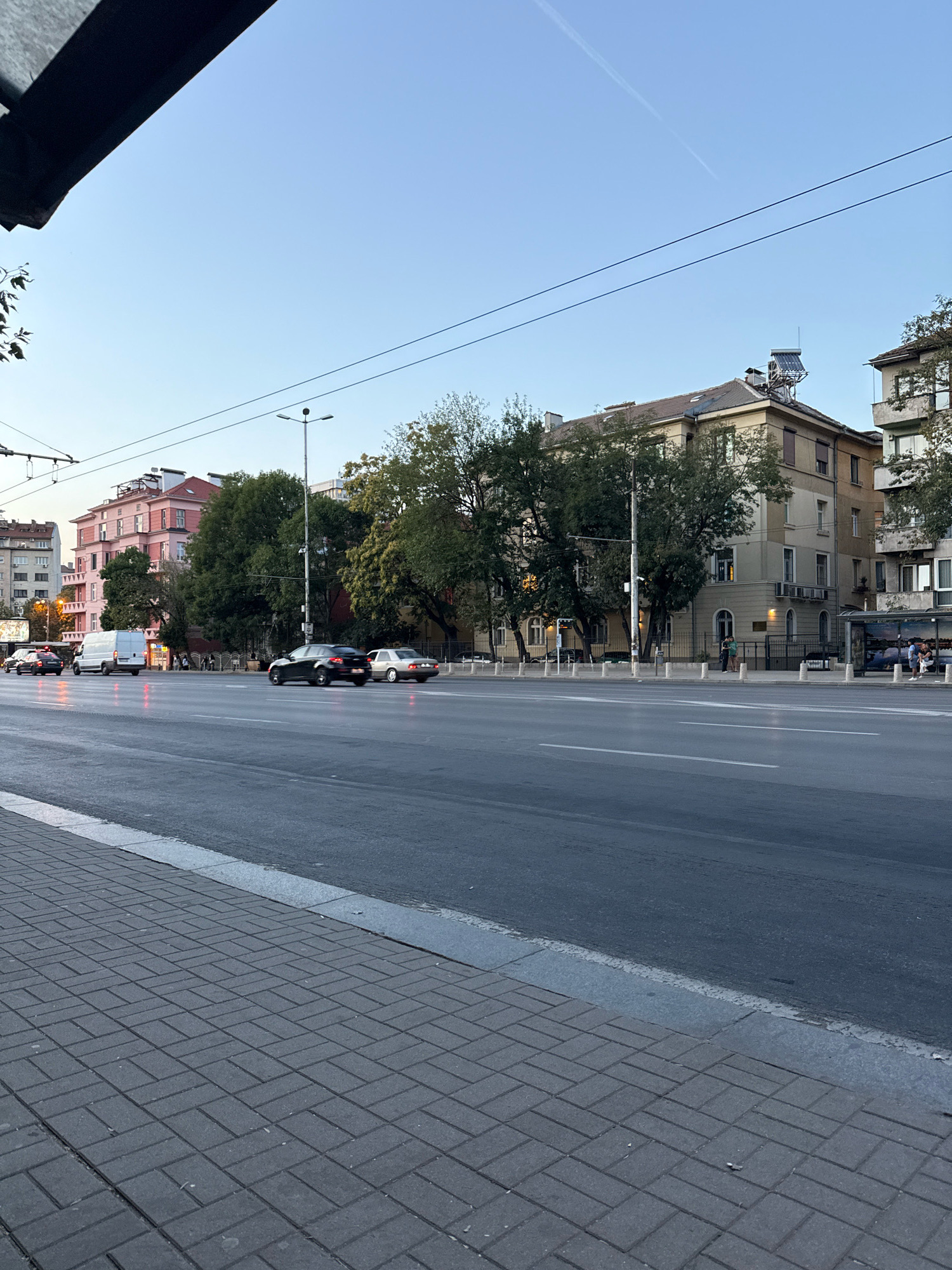 The image captures a street scene in Sofia, Bulgaria, likely during late afternoon or early evening. A wide, multi-lane road dominates the foreground and midground, with asphalt showing some reflectivity, suggesting recent rain or dampness. Paving stones form a sidewalk along the right edge of the road.
On the far side of the road, a row of buildings lines the street. The most prominent building is a light-colored, multi-story structure with several windows and air conditioning units. To its left, a pink building with white trim is visible. Trees with dense green foliage are interspersed between and in front of the buildings.
Several cars are present on the road: a white van, a black sedan, a silver sedan, and a few other vehicles further in the distance. A bus stop shelter with people waiting inside is visible on the right side of the street. Overhead, power lines and streetlights are strung across the scene. The sky is a clear, pale blue with a faint contrail from an aircraft.
The overall impression is a typical urban street in Sofia, with active traffic and pedestrians, set against a backdrop of residential or commercial buildings.