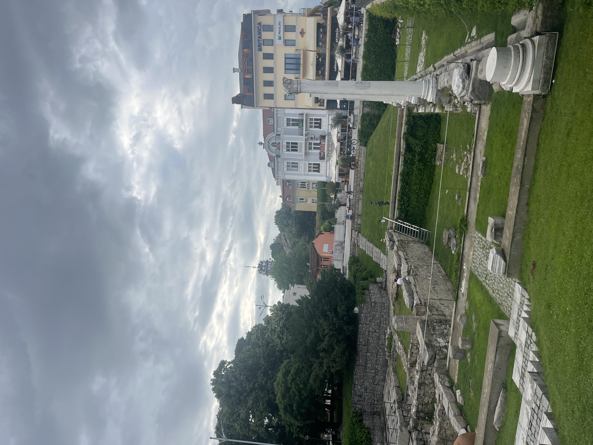The image depicts the Ancient Stadium of Philippopolis in Plovdiv, Bulgaria, viewed from an elevated perspective. The foreground contains the exposed ruins of the stadium, including stone walls, steps, and scattered blocks. A grassy area with remnants of columns and foundations lies adjacent to the ruins. In the background, buildings are visible, including the Britannica building with the MetLife logo and a building with cafes and restaurants. People are present in the background near the buildings, some appearing to be tourists or locals walking or dining. The sky is cloudy and overcast. The vegetation consists of trees and shrubs, adding to the urban landscape.