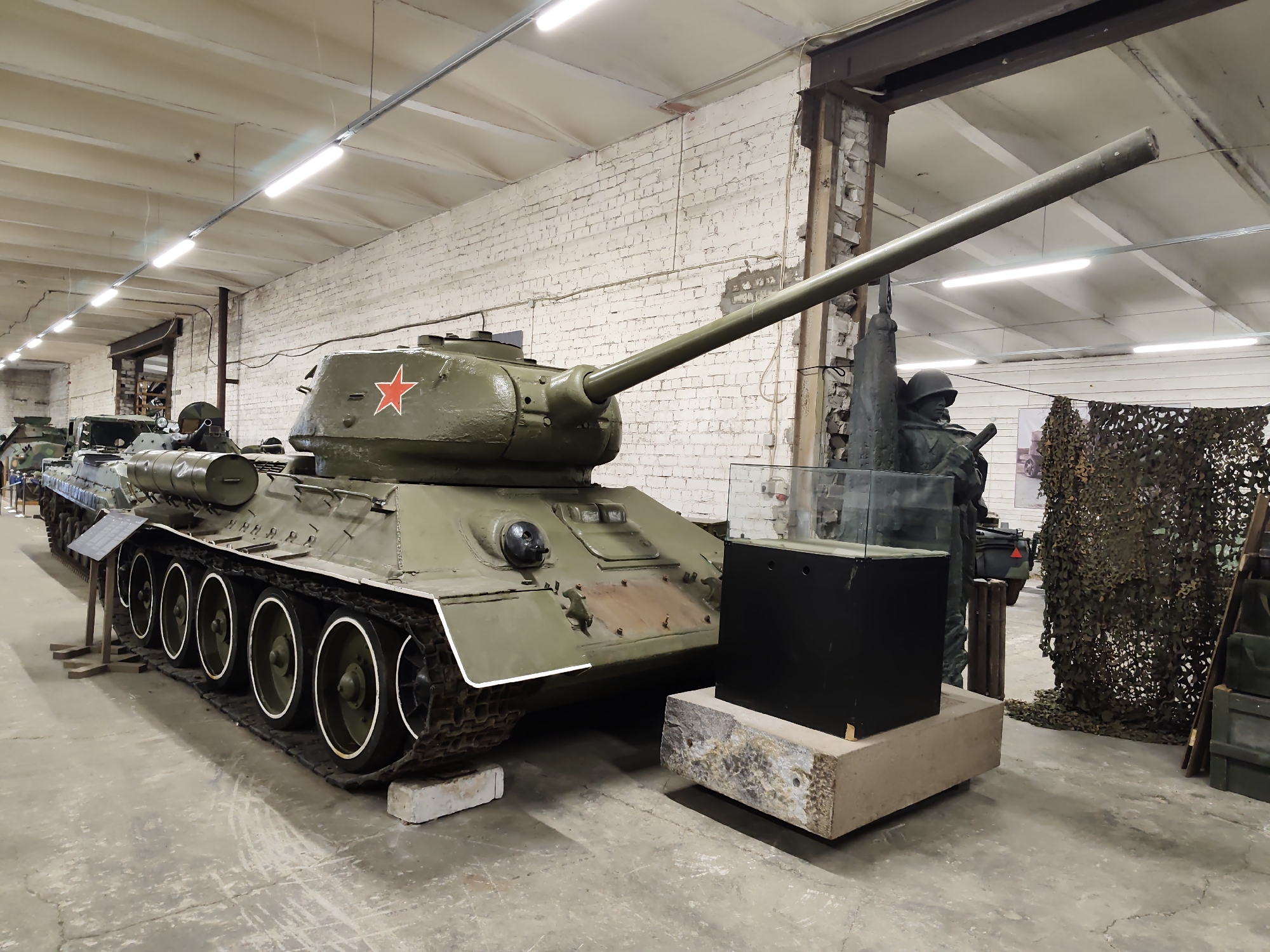 An olive green Soviet T-34 tank is on display in a museum hall, with its barrel angled upwards. The tank has a red star emblem on its turret. In the background, there is a brick wall, fluorescent lighting fixtures, and other military vehicles and equipment. A mannequin of a soldier is visible to the right of the tank, and camouflage netting is draped behind it. The floor is concrete. The image appears to be taken at the Viimsi Open Air Museum in Viimsi, Estonia, which houses a collection of Soviet military equipment. There are no people actively interacting with the tank in the image.