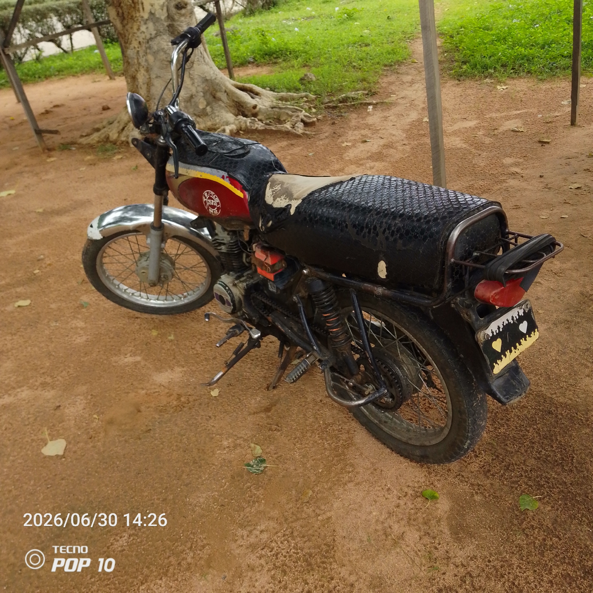 A motorcycle is parked on a dirt path next to a large tree. The motorcycle is red and black, with a worn black seat that has a textured pattern. The front wheel is chrome and silver, and the rear wheel is black. There are two metal poles near the motorcycle, one to the left and one to the right. Behind the motorcycle, there is green grass and the trunk of a large tree with exposed roots. The ground is covered in dry, reddish-brown dirt with some scattered leaves.
Visible text on the image includes a date and time stamp: "2026/06/30 14:26" and "TECNO POP 10". There is also a circular logo with "TECNO" inside and "POP 10" below it in white lettering, and a circular emblem on the side of the motorcycle's fuel tank that appears to be a logo as well. A license plate on the back of the motorcycle is hand-painted with yellow and white accents and two hearts, one yellow and one white.
The scene appears to be outdoors in a natural setting. The lighting suggests it is daytime, possibly late afternoon, with diffuse light indicating an overcast sky or shade. The overall mood is neutral, depicting a parked vehicle in a somewhat rustic environment. The location is identified as Jalingo, Nigeria.