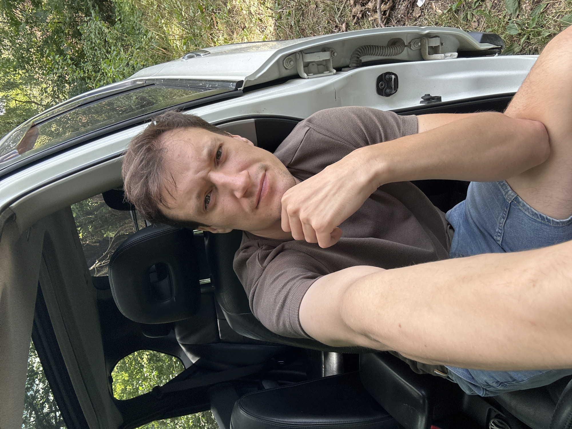 A white man in his 30s with short brown hair and a light brown t-shirt and blue denim shorts is sitting in the driver's seat of a car, which is parked outdoors. The car door is open, and the background is a forest with green trees and foliage. He is looking at the camera and has his right hand resting on his chin, with his left arm extended towards the camera, suggesting he is taking a selfie. The interior of the car has black leather seats and a sunroof. The scene appears to be in the village of Makiv, Ukraine, based on the context provided.