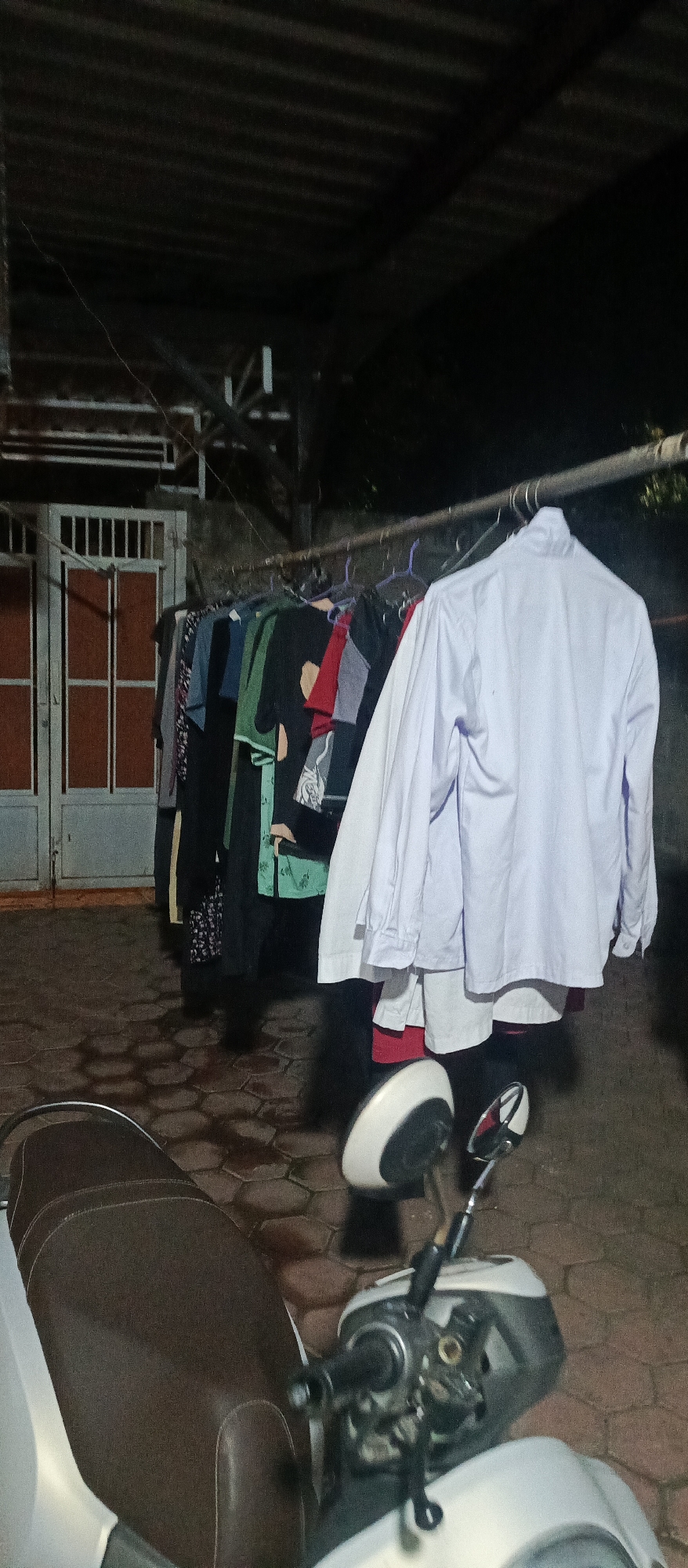 This image depicts an outdoor scene at night, likely in Jatisari, Indonesia, with a clothesline full of laundry hanging from a metal pole. The clothes appear to be a mix of casual wear and possibly some uniforms, including a prominent white shirt hanging on the right side of the frame. In the foreground, a motorcycle is parked, with its brown seat and part of the handlebars visible. Behind the clothesline, a building with a corrugated metal roof and a double door with vertical bars is partially visible. The ground is paved with hexagonal tiles. The overall lighting suggests it is nighttime, with artificial light illuminating the scene. There are no people visible in the image.