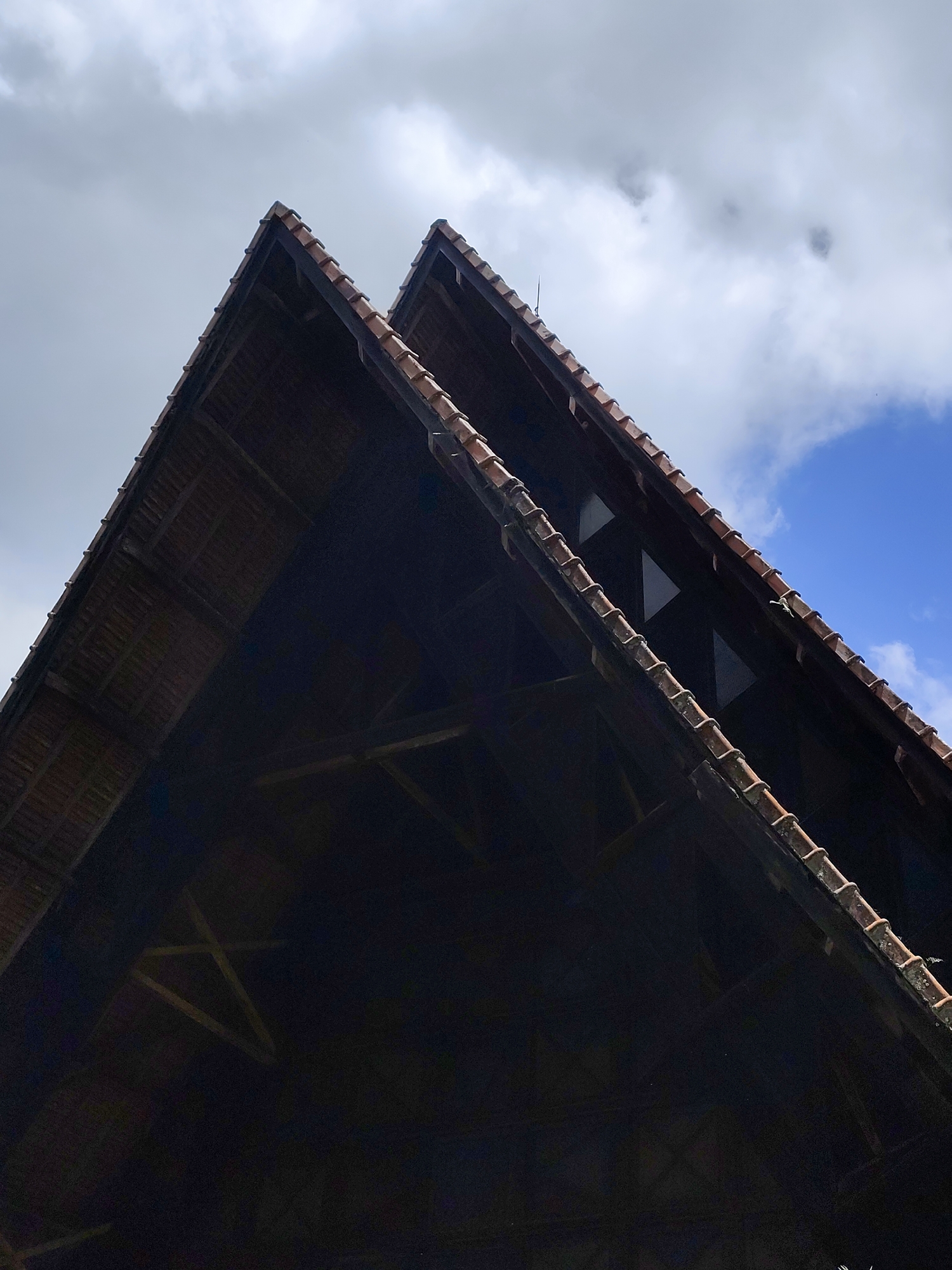 This is a low-angle, close-up, exterior, waist-up shot of two adjacent steeply pitched, dark brown, wooden roof gables, both covered with reddish-brown clay tiles. The two gables meet at a sharp angle, with the left gable slightly taller and positioned in front of the right one. The interior wooden structure of both gables is visible, showing a lattice of crossbeams and rafters. The sky above is a mix of dark, brooding clouds and patches of bright blue sky. The architectural style and material suggest a rustic, possibly historical building, which is consistent with the common styles found in Da Lat, Vietnam, known for its French colonial architecture and pine forests. The image does not contain any people or animals.