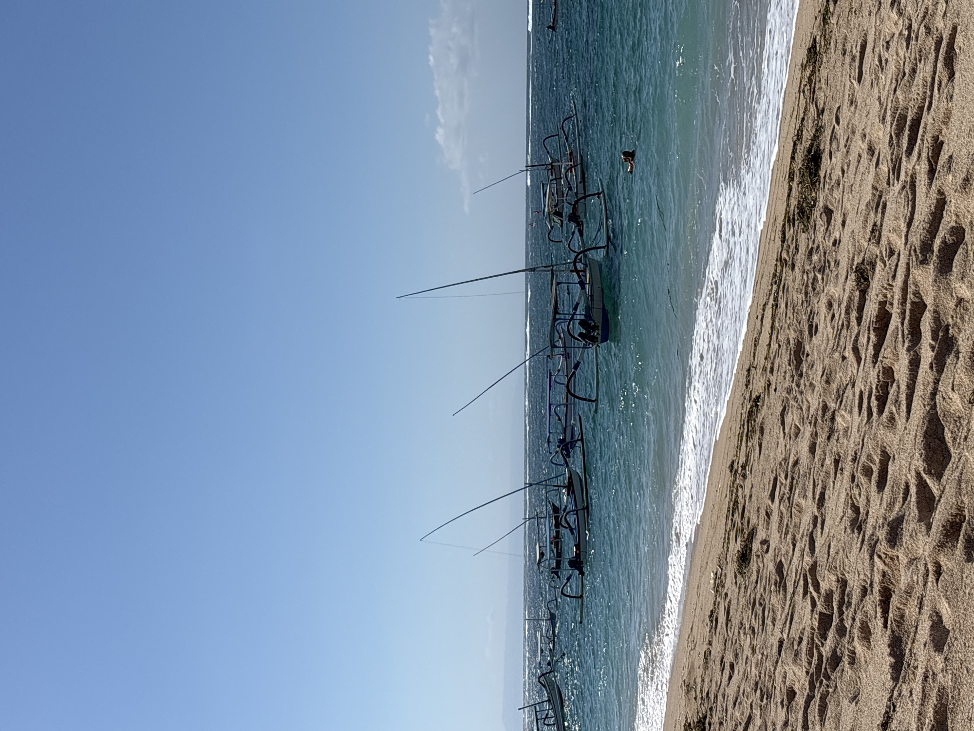The image presents a serene tropical beach scene, likely set in Kuta Selatan, Indonesia, under a clear, bright blue sky, indicating morning or early afternoon. The composition is divided into three horizontal elements: the sky, the ocean, and the beach.
The upper portion of the image features a vast, light blue sky with only a few faint, wispy clouds visible near the horizon. The middle section showcases the sparkling turquoise waters of the ocean, where several traditional Indonesian outrigger fishing boats, known as Jukungs, are gently floating. These boats, distinctive with their slender hulls and extended outriggers, are lined up in the shallow water parallel to the shoreline, their dark silhouettes contrasting against the luminous sea. A solitary individual is partially submerged in the water, with their head and shoulders visible, suggesting they are swimming or wading close to one of the boats. Gentle white-capped waves can be seen breaking along the shore.
The lower part of the image displays the sandy beach, which is light brown and textured with distinct ripple patterns, possibly created by tides or footsteps. Sparse patches of dark green foliage or seaweed are scattered along the sand near the water's edge. The overall impression is one of peacefulness and natural beauty. No visible text is present in the image.