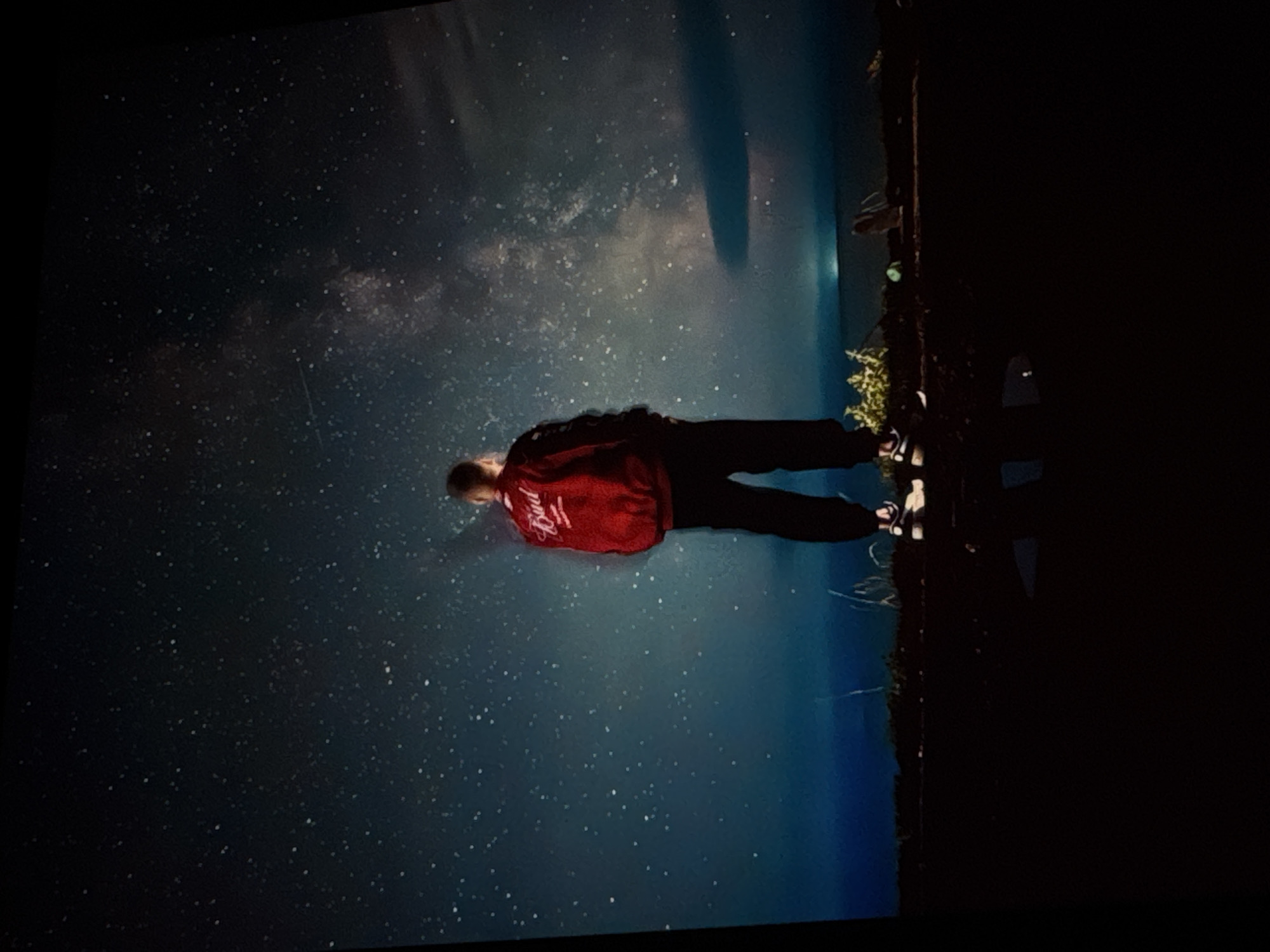 A person stands facing away from the camera, gazing at the Milky Way. The person is wearing a red jacket with "Bud" embroidered on the back and black pants. They are standing on uneven, dark terrain, possibly a hilltop or embankment, with some sparse vegetation visible at the base. The sky is a deep blue, filled with countless stars and a prominent band of the Milky Way galaxy stretching across it. In the distance, a faint, hazy light suggests the presence of a distant city or artificial light source on the horizon. The overall atmosphere is one of awe and contemplation of the night sky. There is no visible indication that this scene is specifically located in Irkutsk, Russia.