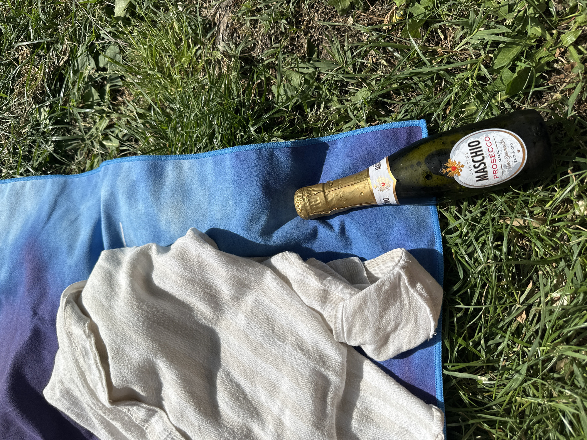 A partially-unfolded white linen shirt rests on a blue and purple ombre towel spread on a patch of green grass. A bottle of Maschio Prosecco lies next to the towel, partly on the grass and partly on the towel. The scene appears to be outdoors, possibly in a park or garden.