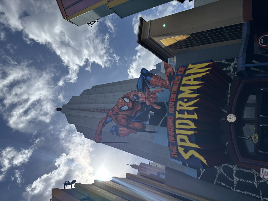 The image displays the exterior of "The Amazing Adventures of Spider-Man" attraction at Universal's Islands of Adventure in Orlando, Florida, United States of America. A large, three-dimensional representation of Spider-Man in his classic red and blue suit is prominently featured, crouching and extending his left hand forward, positioned above the attraction's primary entrance. Below this figure, the ride's title, "THE AMAZING ADVENTURES OF SPIDER-MAN," is displayed in yellow gothic-style lettering on a dark blue and red sign.
Behind the Spider-Man figure stands a tall, light-colored Art Deco-style skyscraper, representative of the Daily Bugle building, with "DAILY BUGLE" inscribed vertically on its facade. Additional stylized buildings, consistent with a comic book urban setting, are visible on both the left and right sides of the frame. The lower portion of the entrance features a dark blue facade with arched doorways, one of which displays an "EXPRESS" sign. A small, circular white clock with a red rim is mounted above the central entrance arch. The scene is illuminated by bright daylight under a partially cloudy blue sky, with the sun's glare visible on the upper left portion of the image. No individuals are visible in the frame.