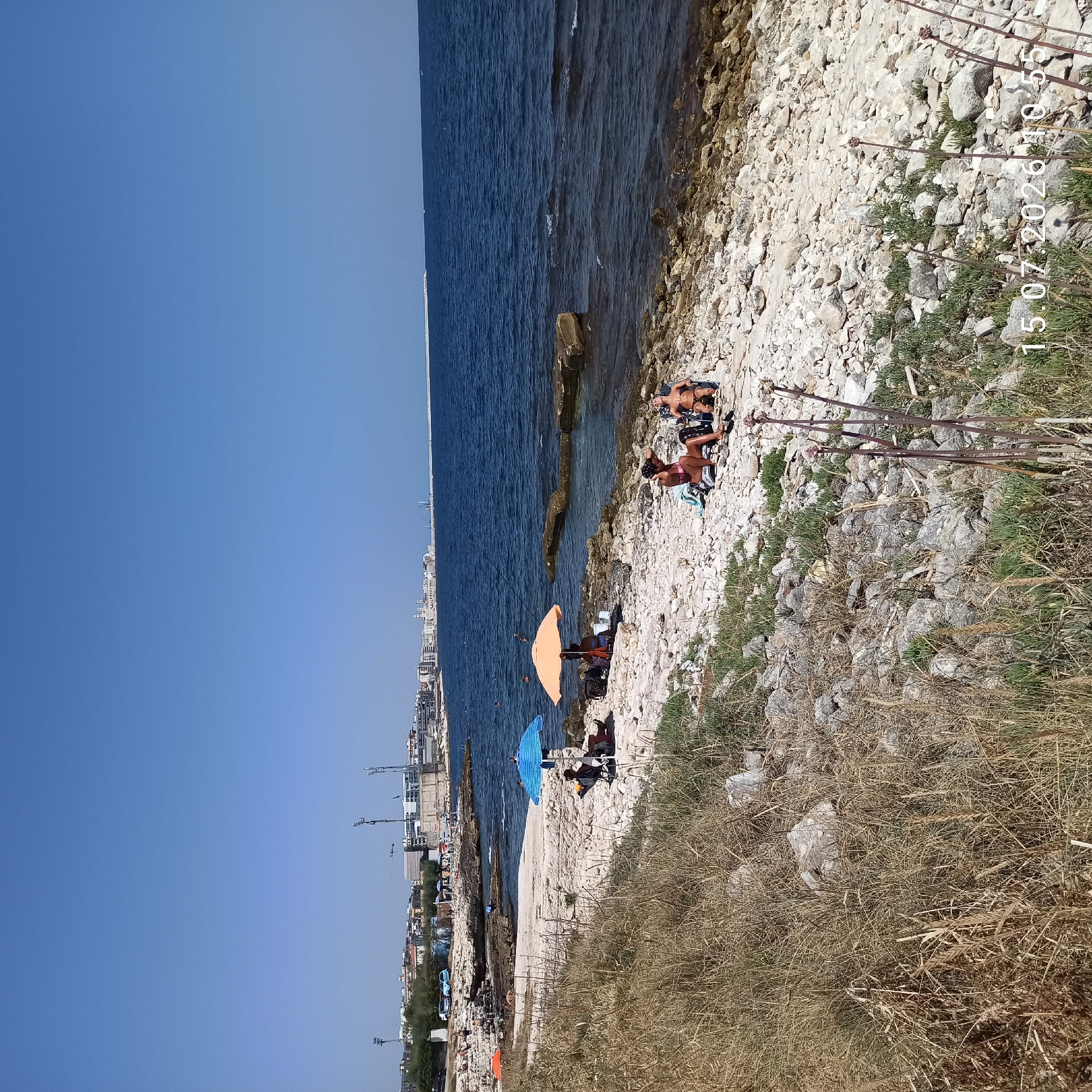 The image captures a sunny day at a rocky beach in Molfetta, Italy, with the city skyline visible in the background. The foreground features dry grass and scattered rocks. In the middle ground, several people are relaxing on the rocky shore. Two individuals are seated under a blue and white striped umbrella, while another pair is under an orange umbrella. Further down the beach, two women are sunbathing in chairs, one wearing a bikini and the other a swimsuit. The sea is a deep blue, with some gentle waves lapping against the shore. The city skyline includes buildings, cranes, and what appear to be stadium lights. The timestamp "15.07.2026 10.55" is visible in the lower right corner, indicating the date and time the photo was taken.