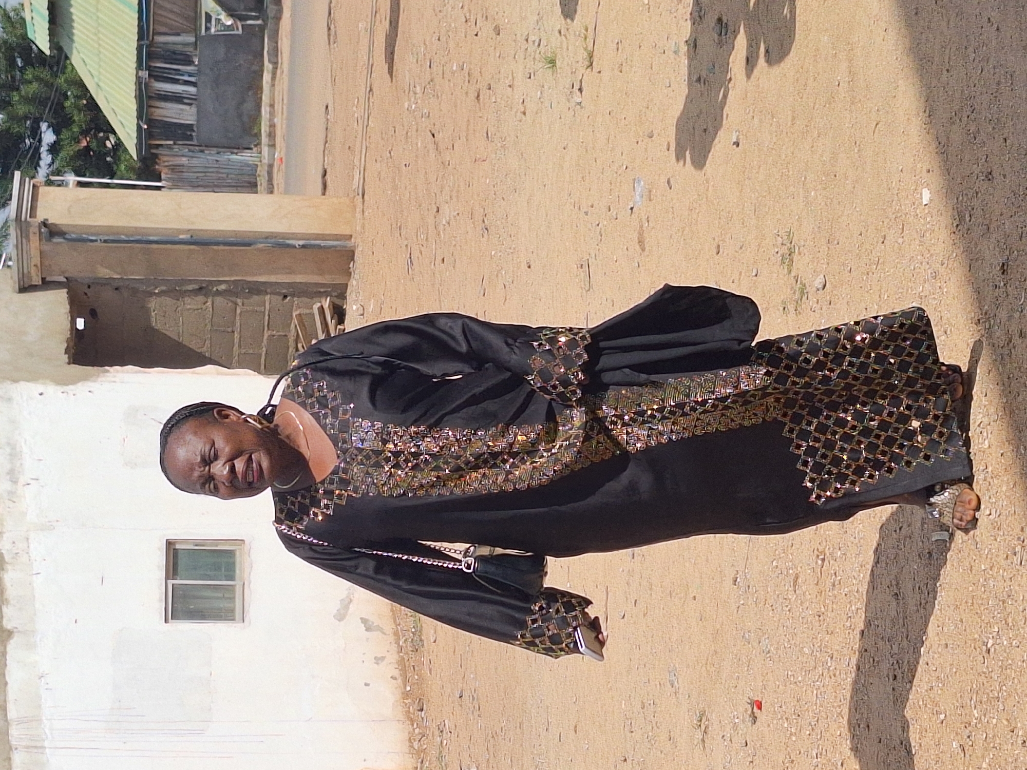 The image shows a woman standing in Sabon Tasha, Nigeria. She is wearing a long, black dress with gold and black geometric patterns. She has a purse slung over her shoulder and is holding a phone. The background consists of a light-colored building with a window and a sandy ground with shadows.