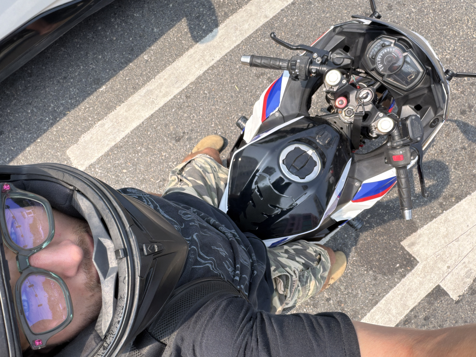 A person wearing a full-face helmet with sunglasses is seated on a motorcycle on a paved surface marked with white lines. The motorcycle is predominantly black, white, and blue, featuring a visible speedometer. The person wears a black shirt and camouflage pants, with tan-colored boots visible near the motorcycle's components. The environment appears to be an outdoor setting during daytime. No evidence indicates the location is within the city of Đà Nẵng, Vietnam.