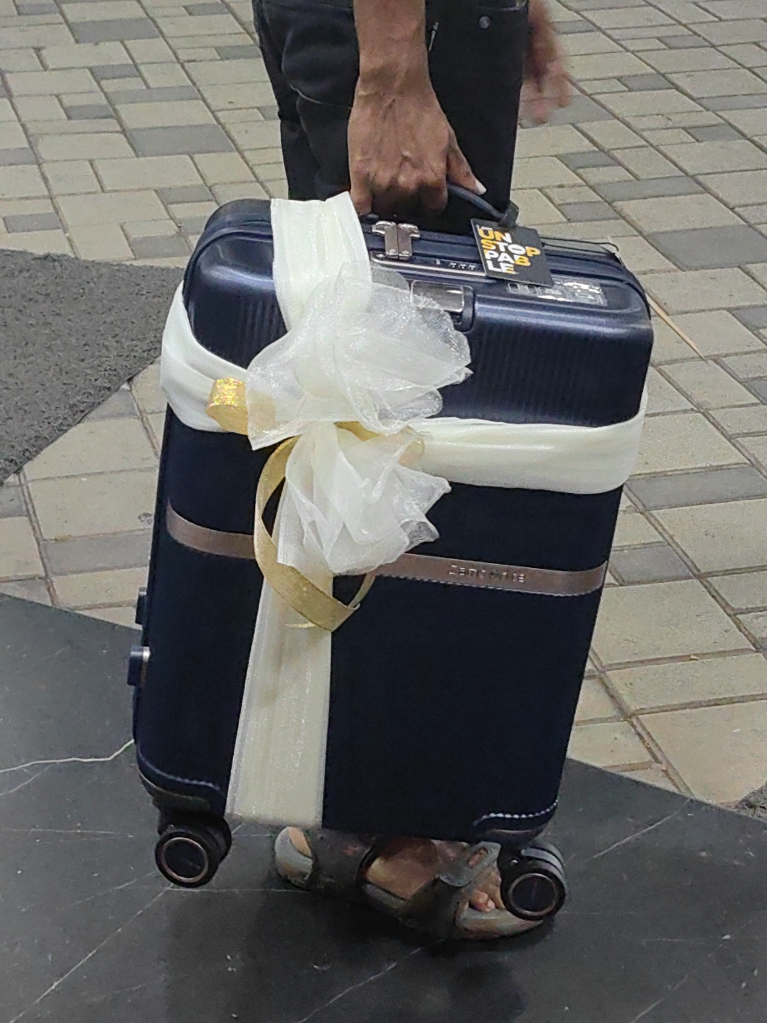 A person's hands hold the handle of a dark blue Samsonite suitcase, which is decorated with white and gold ribbon. The suitcase has a tag attached to it that reads "UNSTOPPABLE". The person is wearing dark pants and sandals. The suitcase is on wheels, with one wheel visible on the right side of the image. The background shows a tiled walkway and a darker tiled surface. The image is taken from a slightly elevated angle, looking down at the suitcase and the person's legs and feet. The scene appears to be outdoors, possibly in a public area like an airport or train station.
