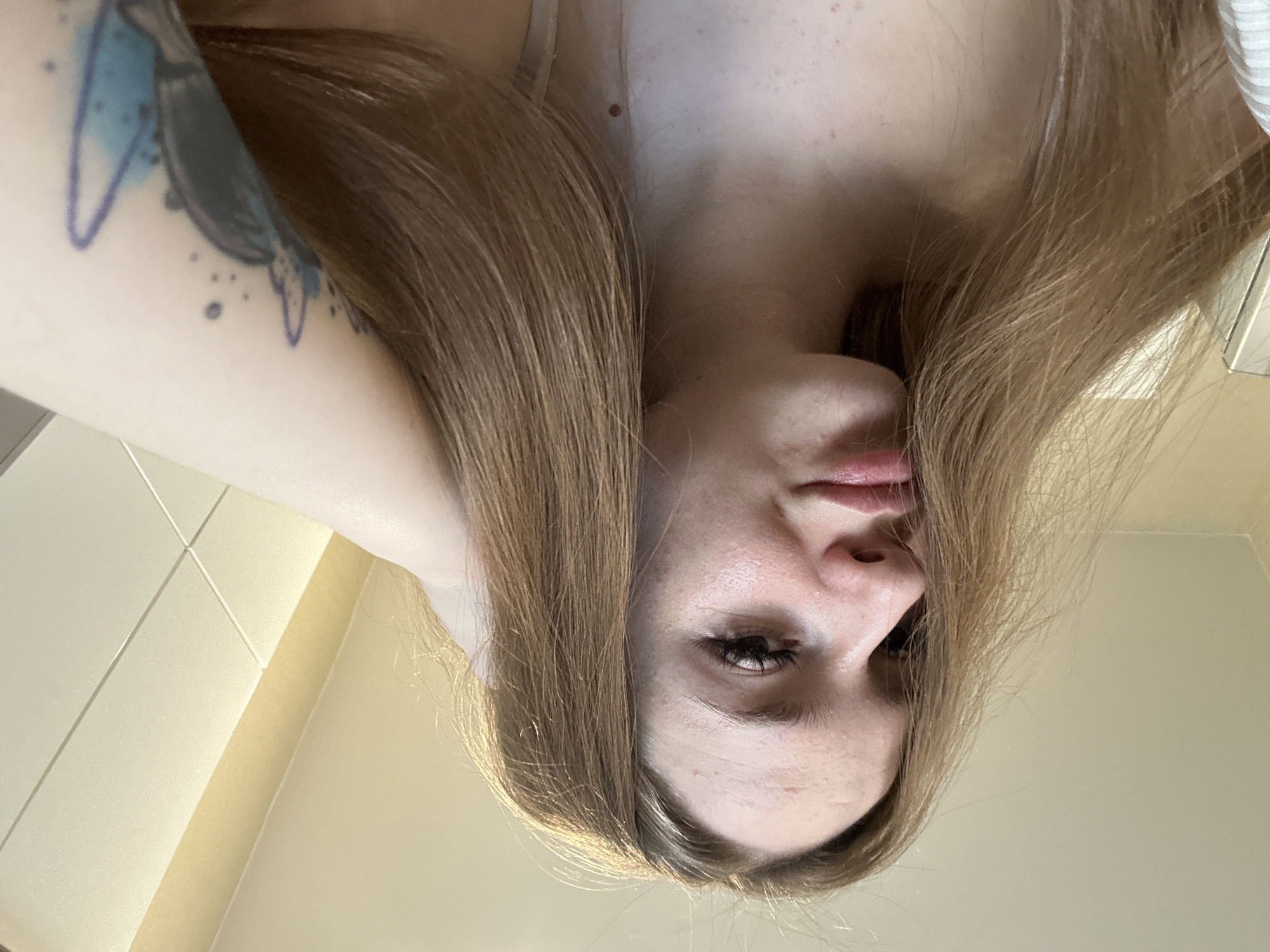 A close-up, upside-down portrait features a young woman with long, light brown hair. Her face is centered in the lower portion of the frame, gazing directly at the viewer from an unusual upward-looking angle. Her hair, parted down the middle, cascades around her face.
Her visible right eye has prominent, dark eyelashes. Her lips are slightly parted, and her skin, which appears fair, shows some minor freckles or blemishes on her forehead and cheeks. On her left arm, a tattoo is visible, featuring light blue abstract shapes reminiscent of watercolor splashes, combined with darker, possibly black or dark blue, linear elements that could suggest a bird or a stylized brushstroke.
A thin, light-colored strap, likely from a tank top or bra, is visible over her right shoulder/chest area, and a small section of ribbed white fabric, possibly clothing, appears on the far right. The background consists of plain, light-colored walls, suggesting an indoor setting. The lighting is soft and diffused. No text is visible in the image.