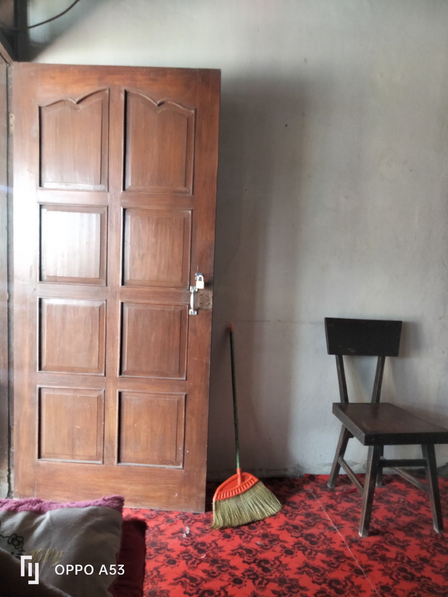 A room in Jati, Indonesia, contains a wooden door, a wooden chair, and a broom. The door, on the left side of the image, is dark brown and has six panels, with a small silver lock attached to the right side. To the right of the door, a wooden chair with a slatted back and seat is positioned. In front of the chair, a broom with an orange plastic head and yellow bristles lies on its side. The floor is covered with a red carpet featuring a black floral pattern. The wall behind the objects is a light gray color. A pillow with a floral pattern is visible in the lower left corner of the image.